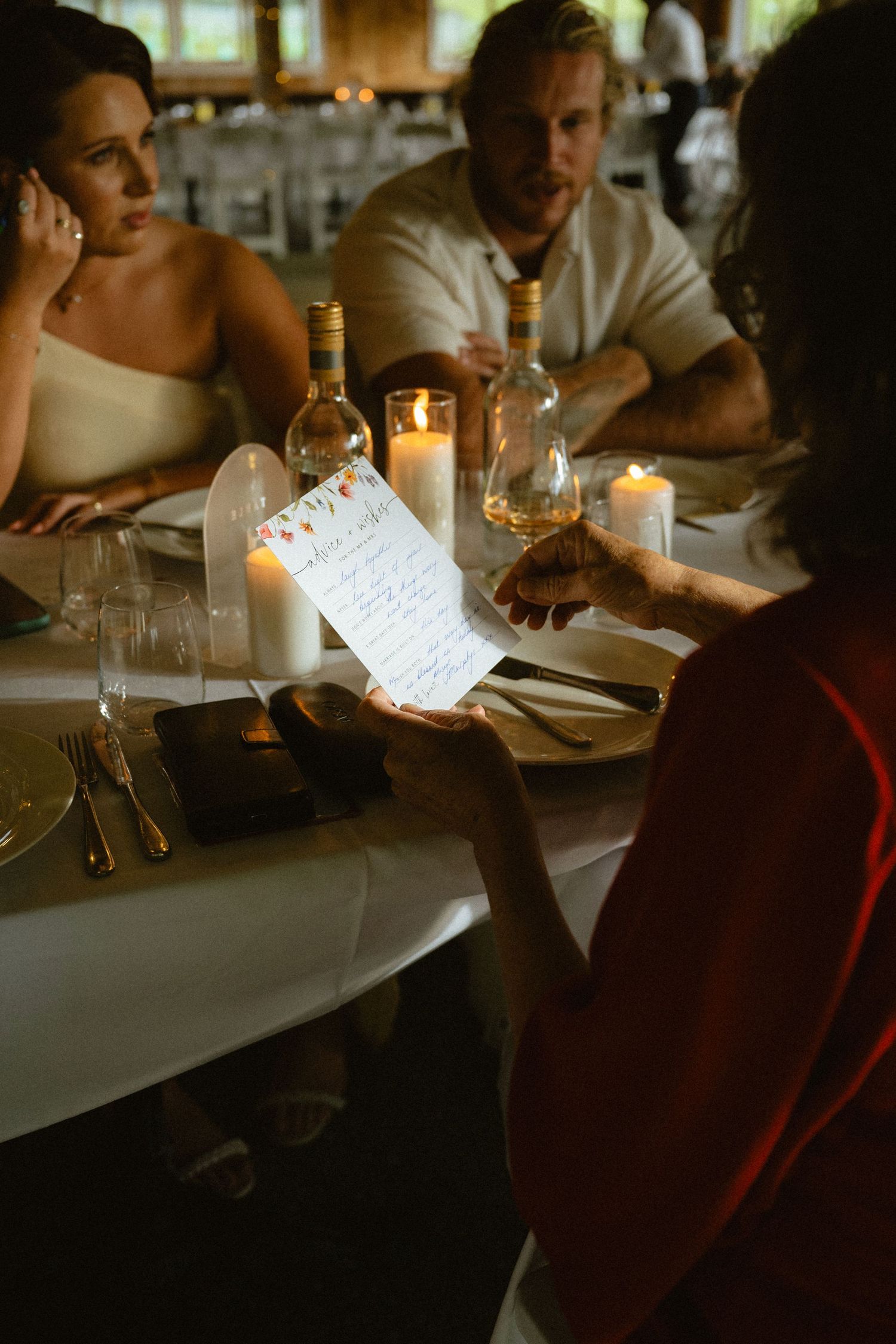 Guests at a candlelit wedding reception table reading menus with wine bottles and glowing candles.