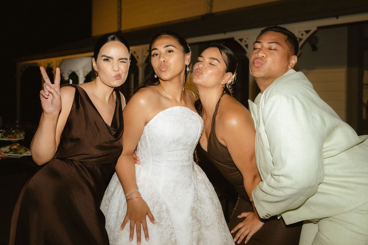 Bride in white strapless gown poses with three bridesmaids in brown dresses making fun faces at wedding reception.