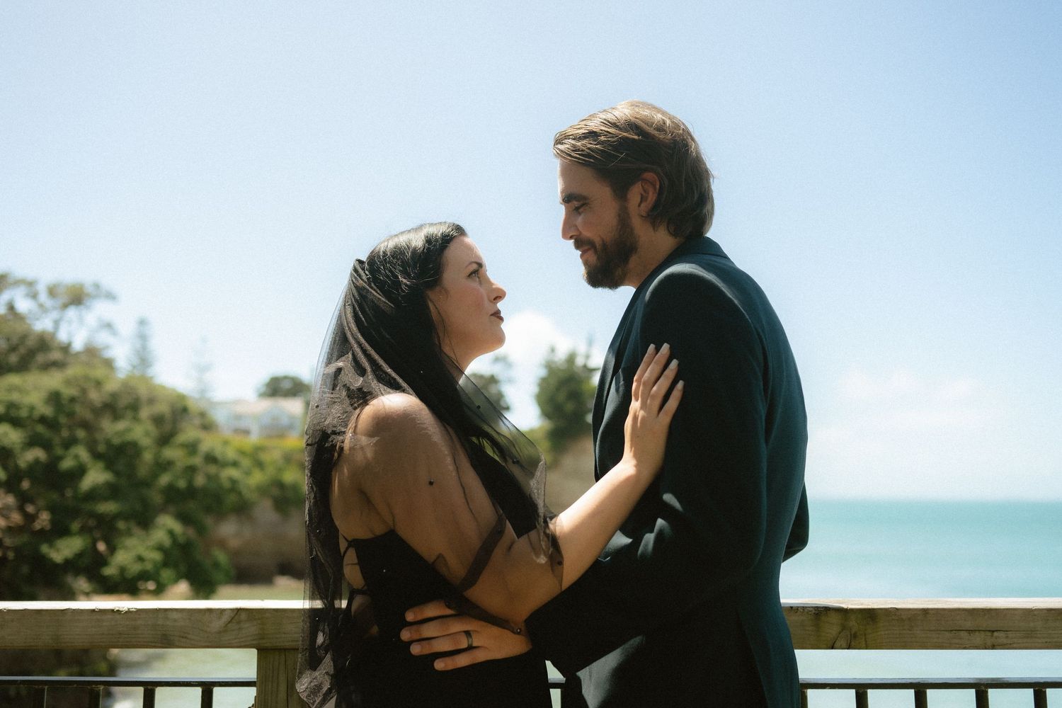 Couple in formal attire sharing a romantic moment on a balcony with ocean views on a sunny day.