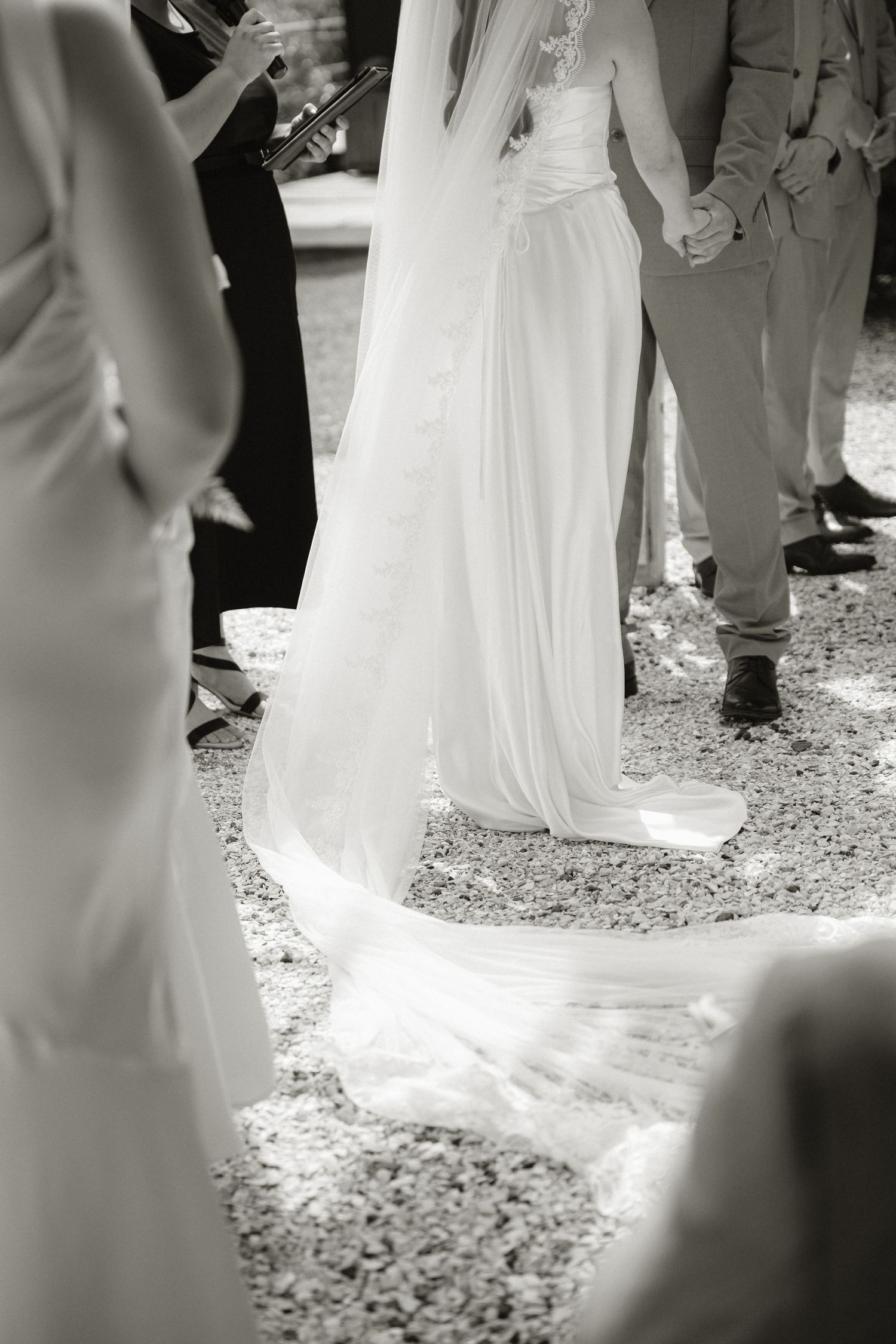 Black and white photo of a bride in a flowing white gown and veil standing with groom during outdoor wedding ceremony.