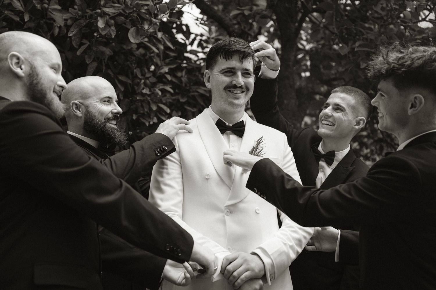 Black and white photo of a groom in white jacket being helped by groomsmen in black suits outdoors.