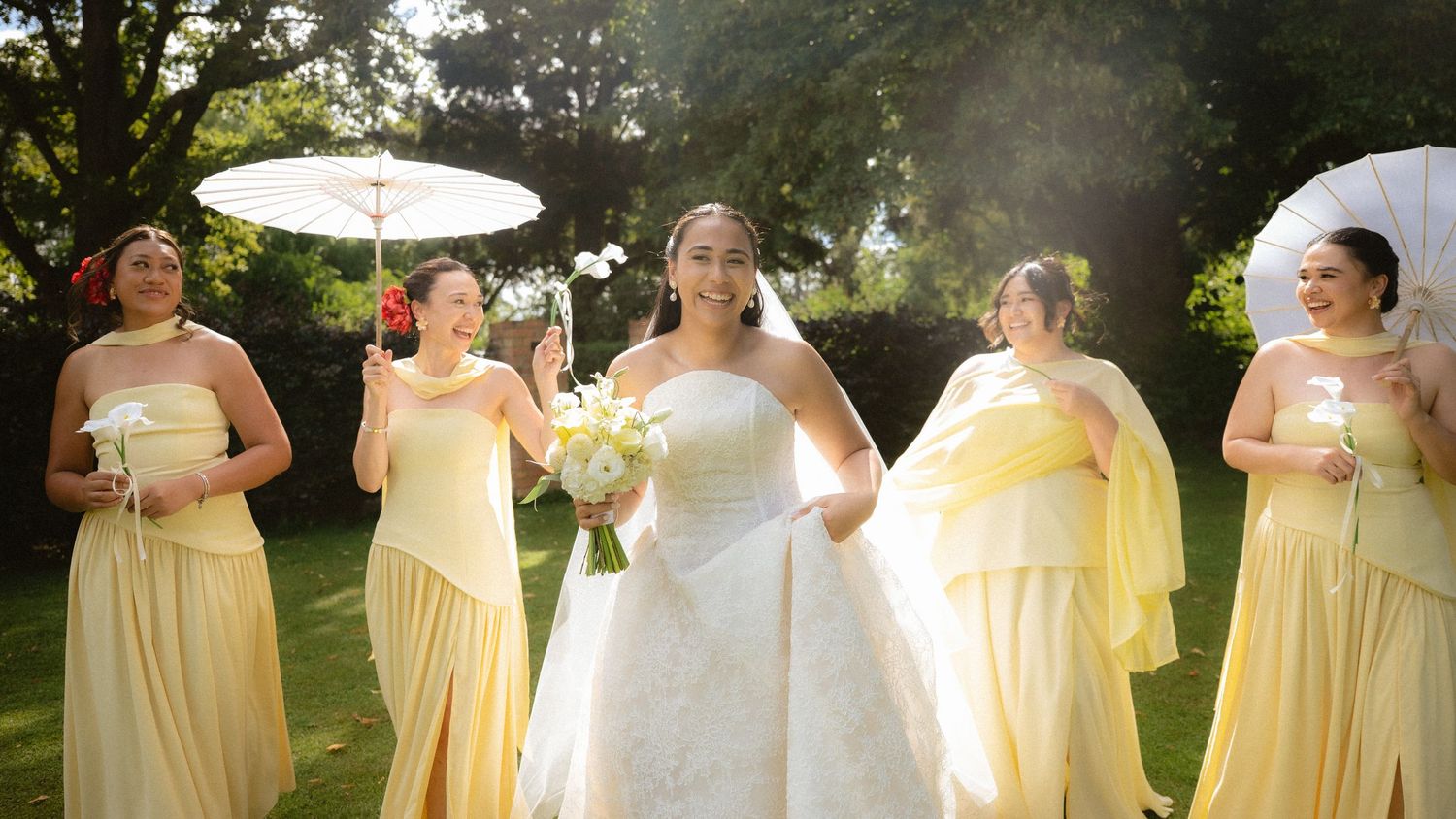 Bride in white gown with bridesmaids in yellow dresses holding parasols outdoors in a sunny garden setting.