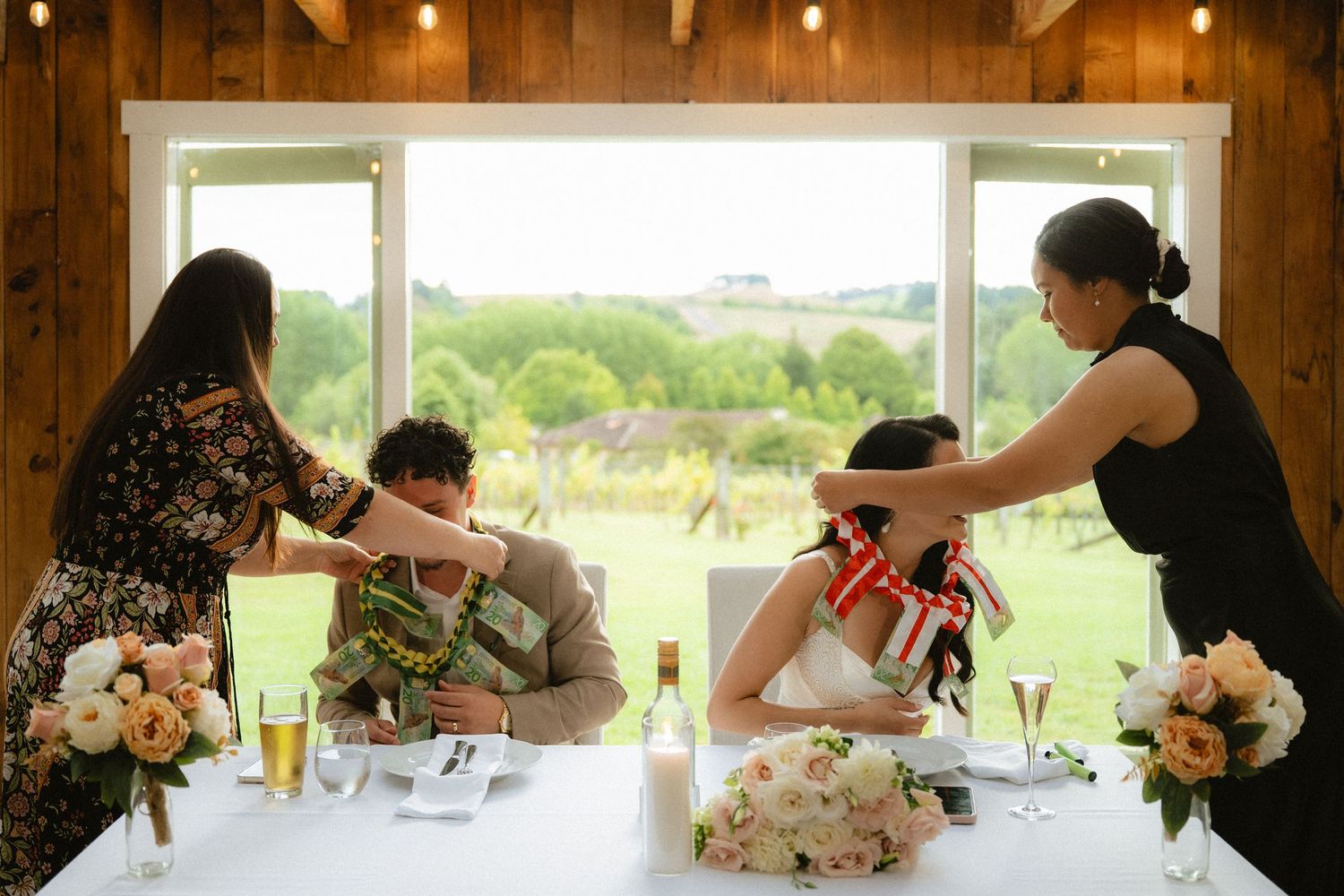 Bride and groom seated at wedding reception table while attendants place leis around their necks indoors.