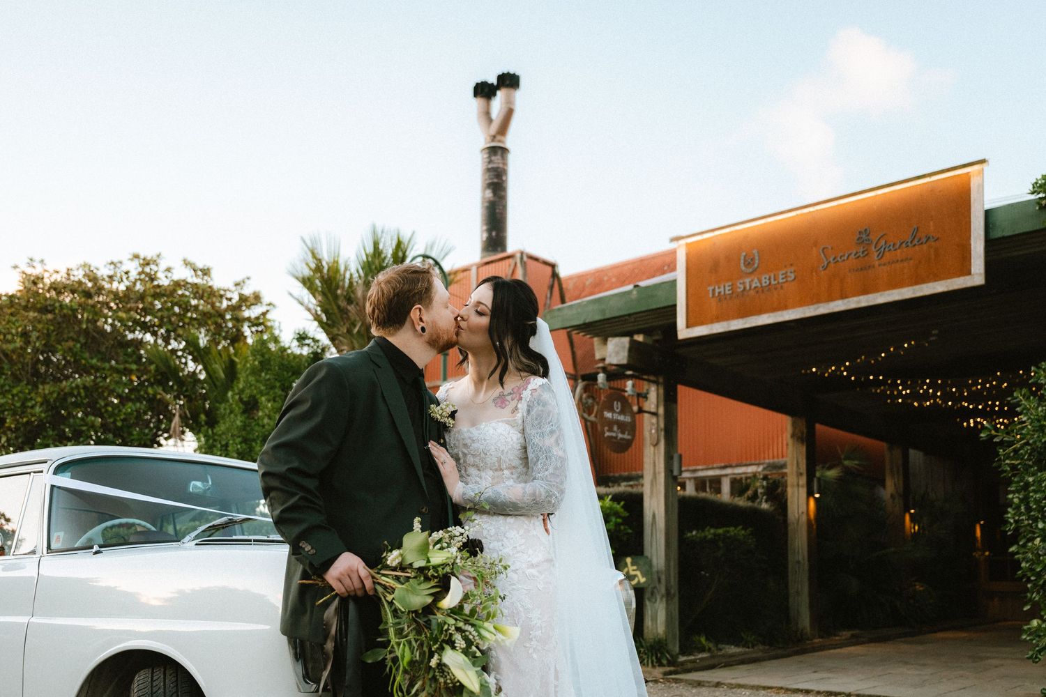 Wedding couple poses outside venue with classic car in background.