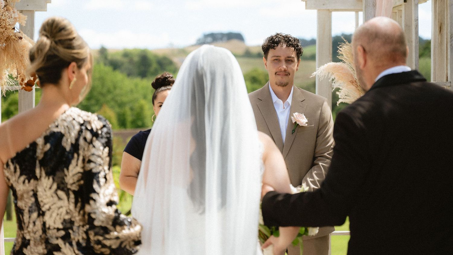 Outdoor wedding ceremony with bride in white veil facing groom in tan suit, surrounded by guests on a green hillside.