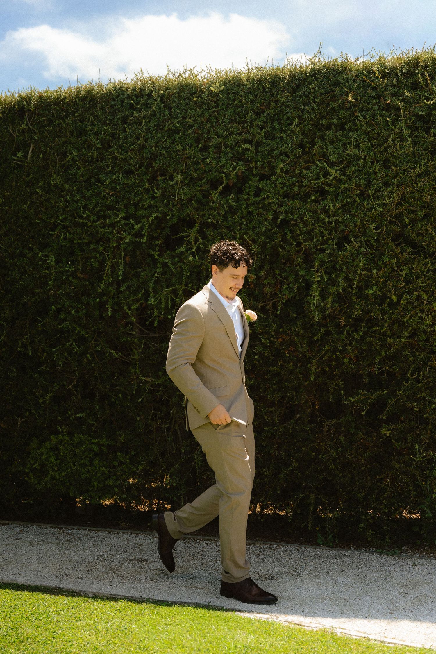 Groom in tan suit with boutonniere walks confidently along garden path beside tall green hedge.