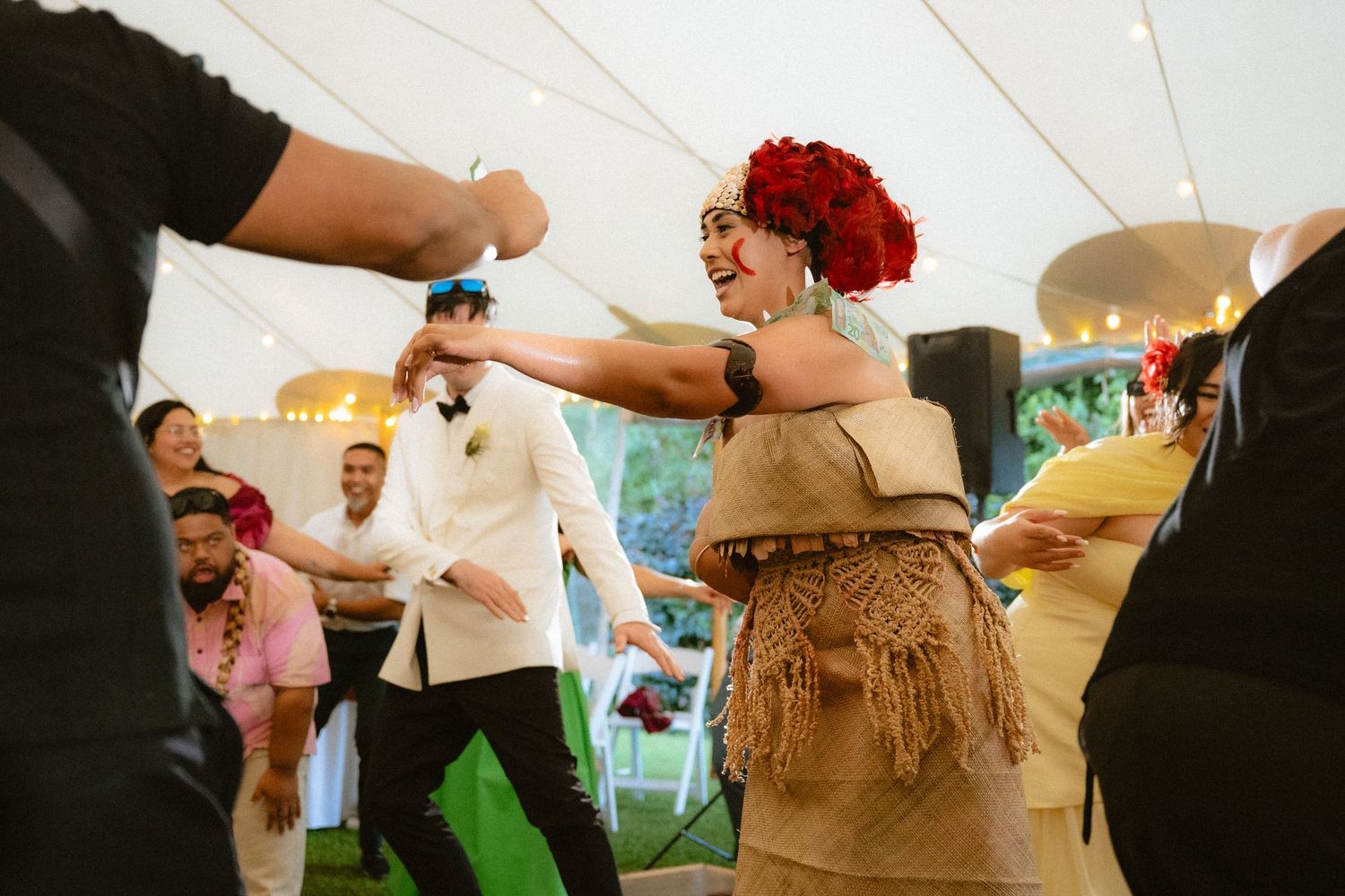 Joyful woman in tan fringe dress and red floral headpiece dancing at a wedding reception under a white tent.