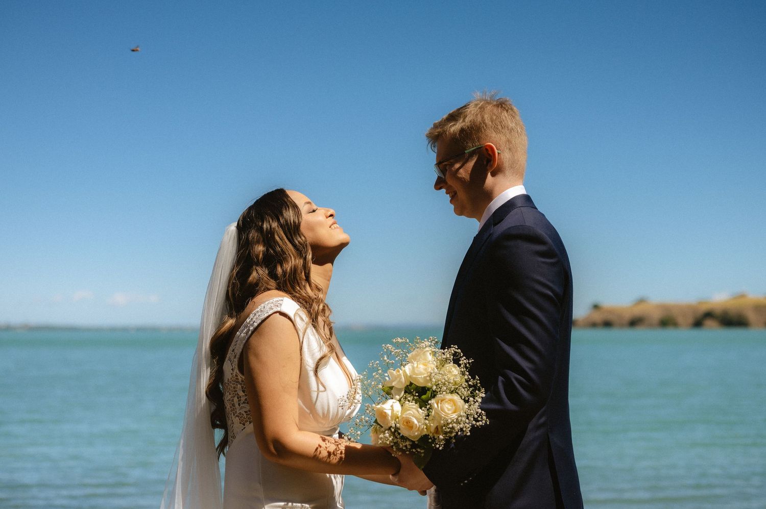 A couple shares an intimate moment on a beach with turquoise waters and blue skies in the background.