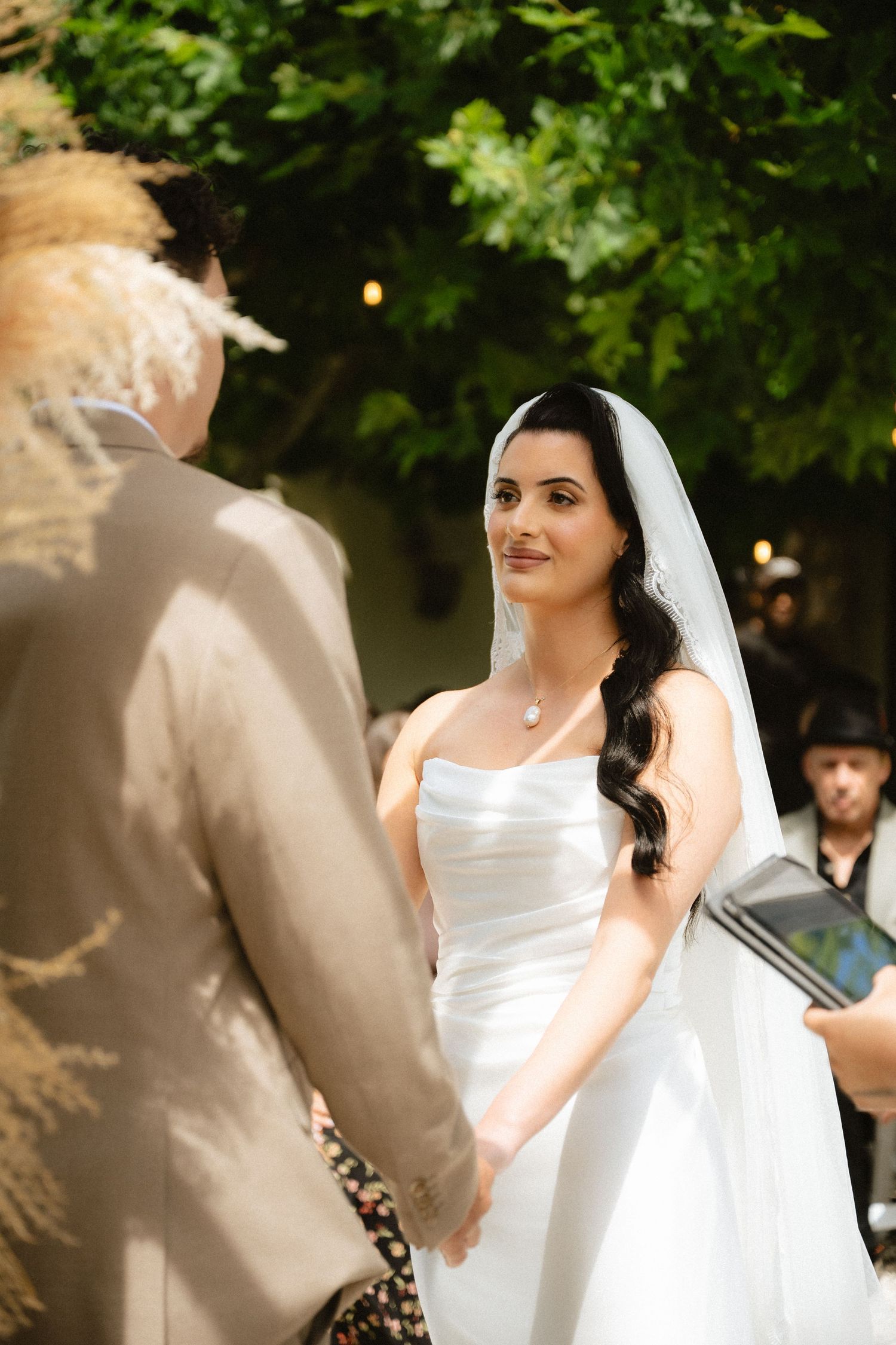 Bride in white strapless gown and veil holds hands with partner during outdoor wedding ceremony under green trees.