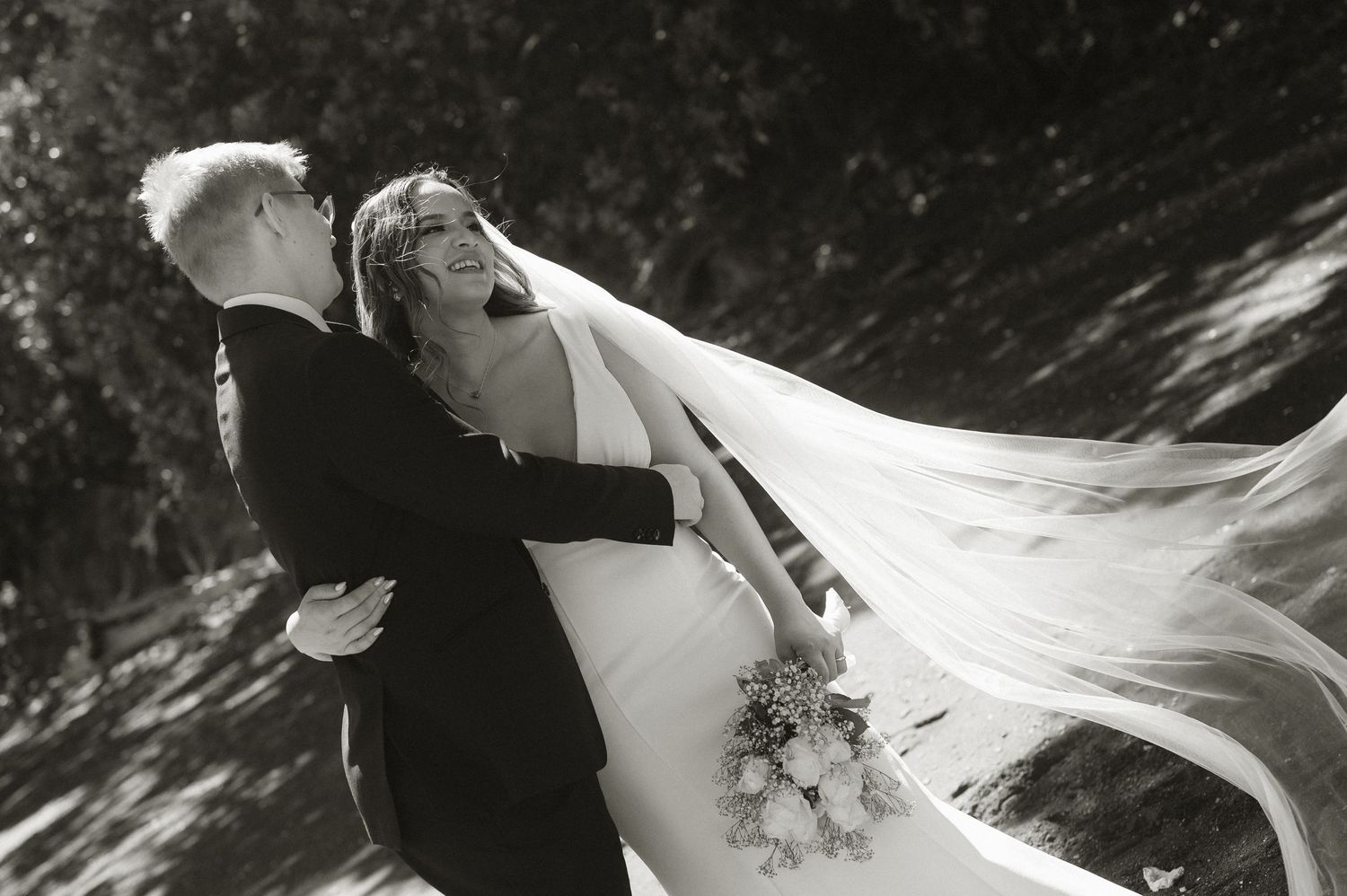 Black and white wedding photo of bride laughing joyfully as groom lifts her, veil flowing in the wind.