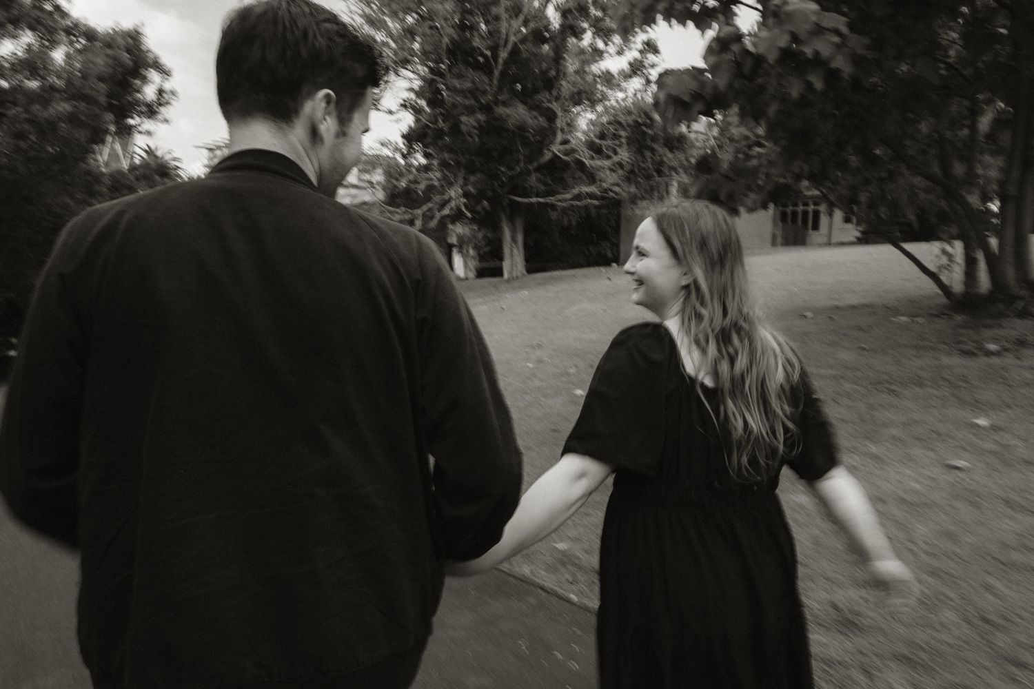 Black and white photo of a couple holding hands, woman in black dress smiling at man as they walk outdoors.