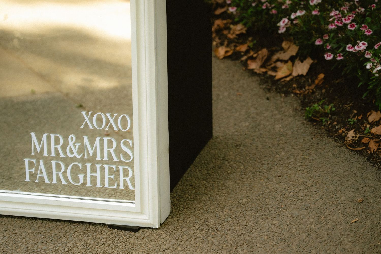White framed mirror with XOXO Mr & Mrs Fargher wedding sign leaning against wall outdoors near pink flowers.