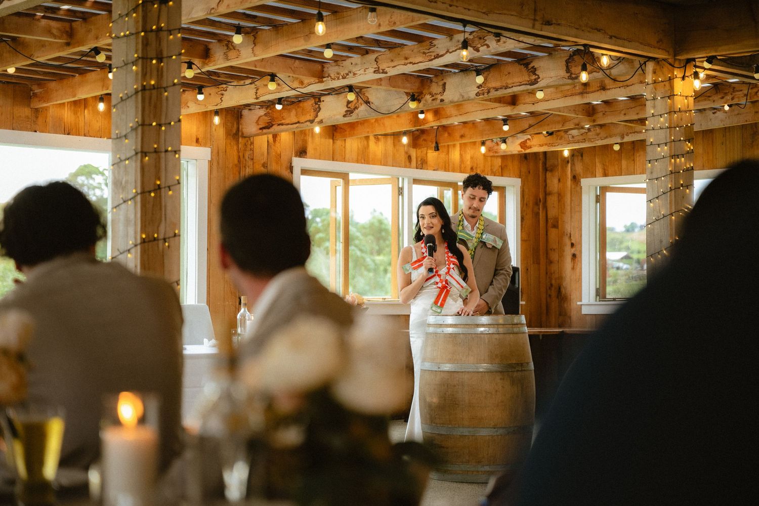 Couple speaking at rustic barn wedding reception with wood walls, string lights, and wine barrel podium.