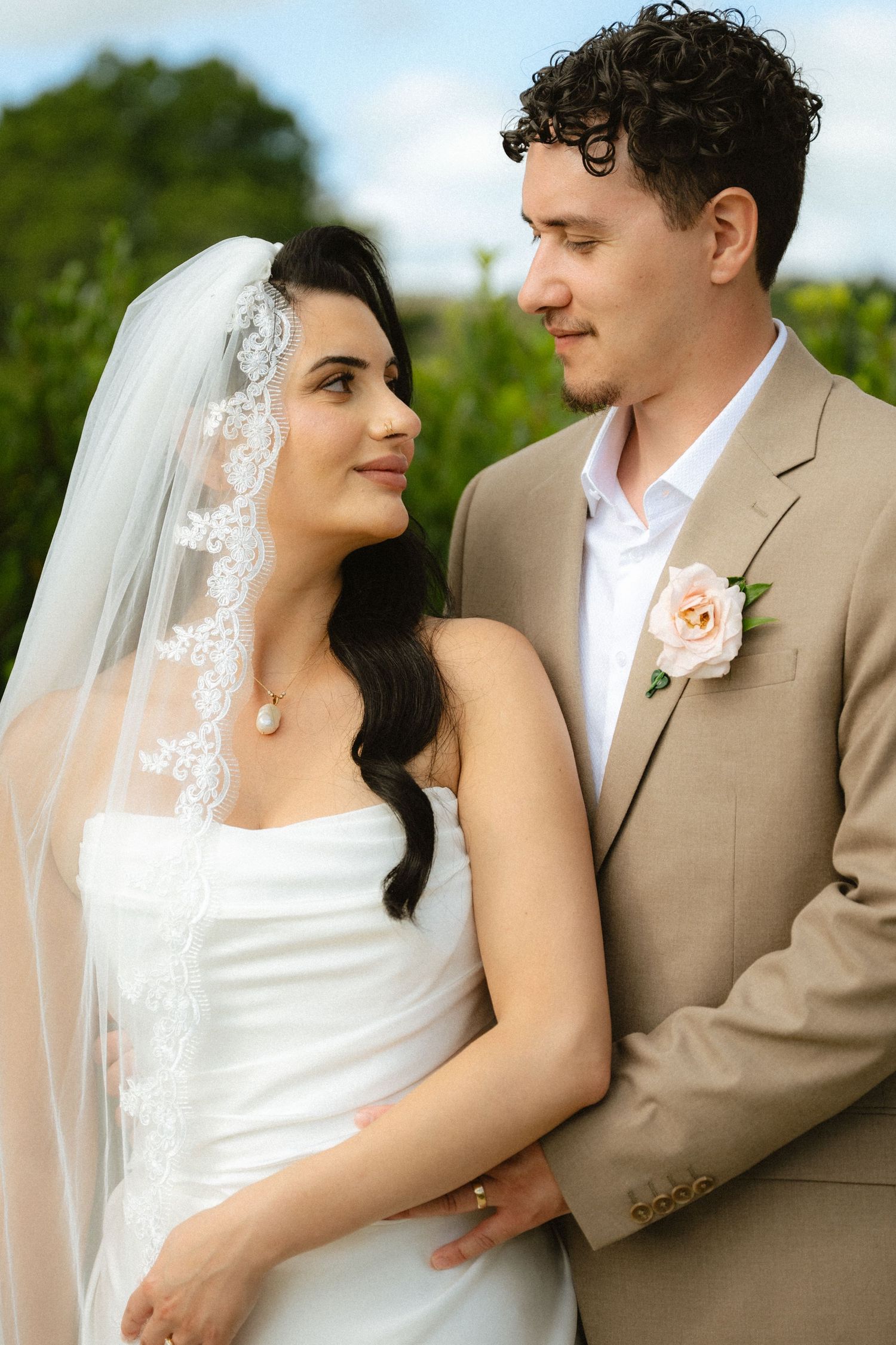 Bride in white gown and lace veil stands with groom in tan suit with pink boutonniere outdoors.