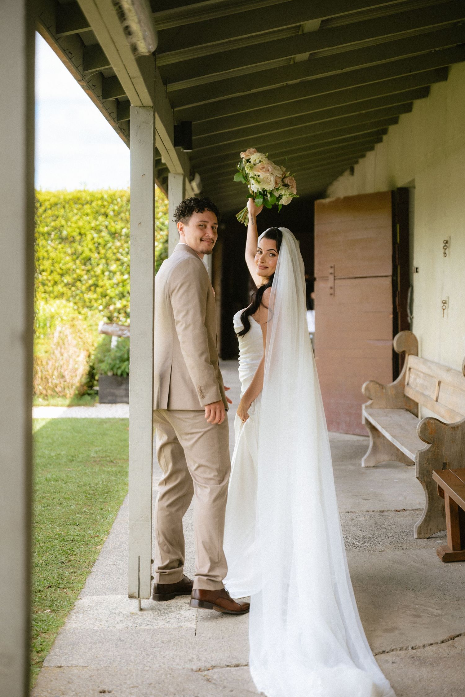 Bride in white gown holding bouquet aloft with groom in tan suit on a covered porch at an outdoor wedding venue.