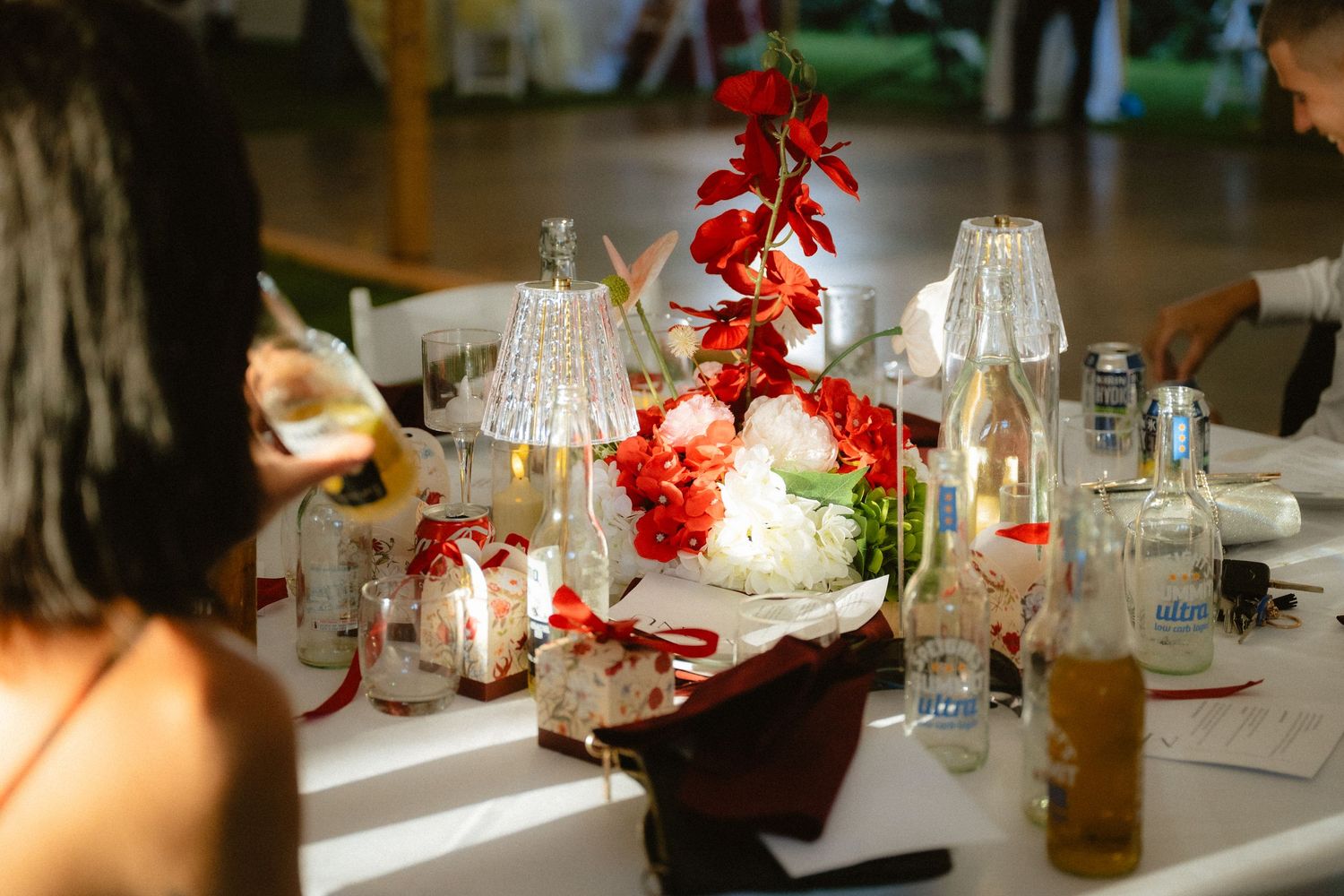 Elegant wedding reception table with red floral centerpiece, white roses, candles, and glassware on a white tablecloth.