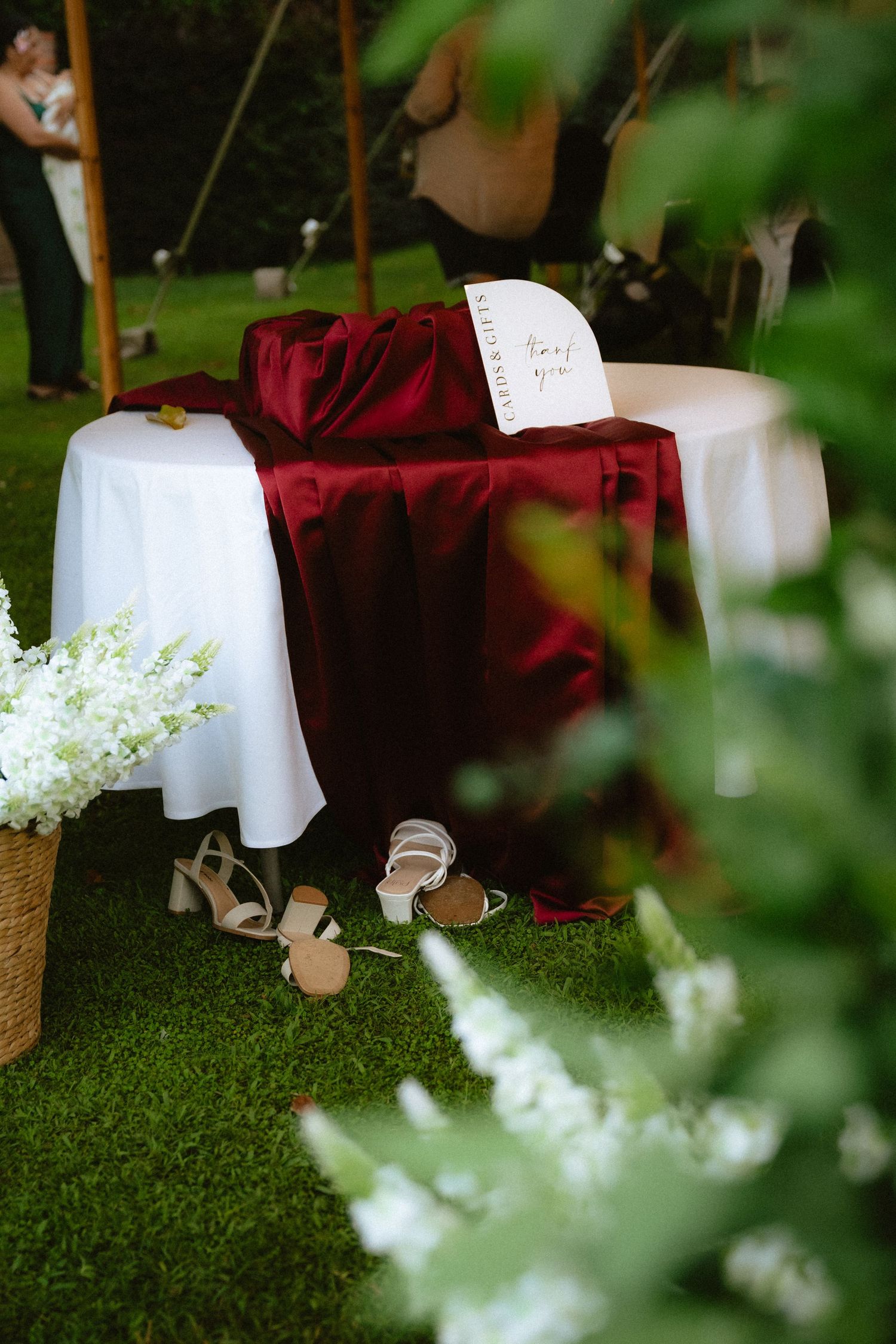Elegant outdoor wedding table with burgundy velvet drape, white florals, and bridal heels on green grass.