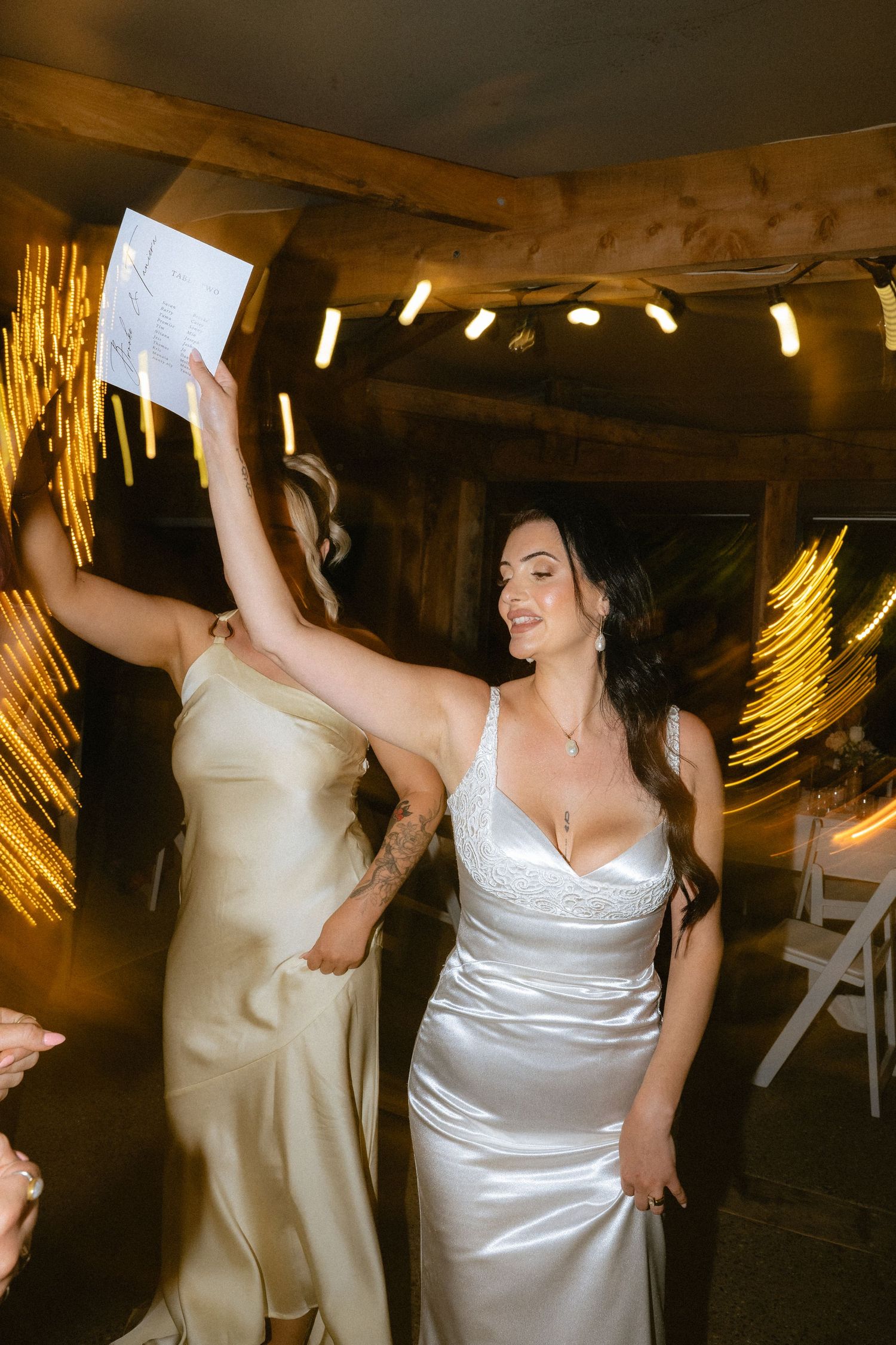 Bride in white satin gown dancing joyfully at wedding reception with bridesmaid in champagne dress, warm rustic venue lighting.