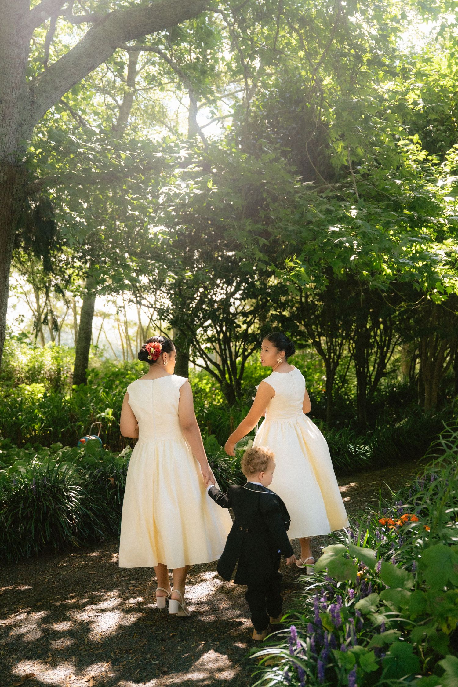 Two flower girls in cream dresses and a ring bearer in black suit walk through a lush garden path.