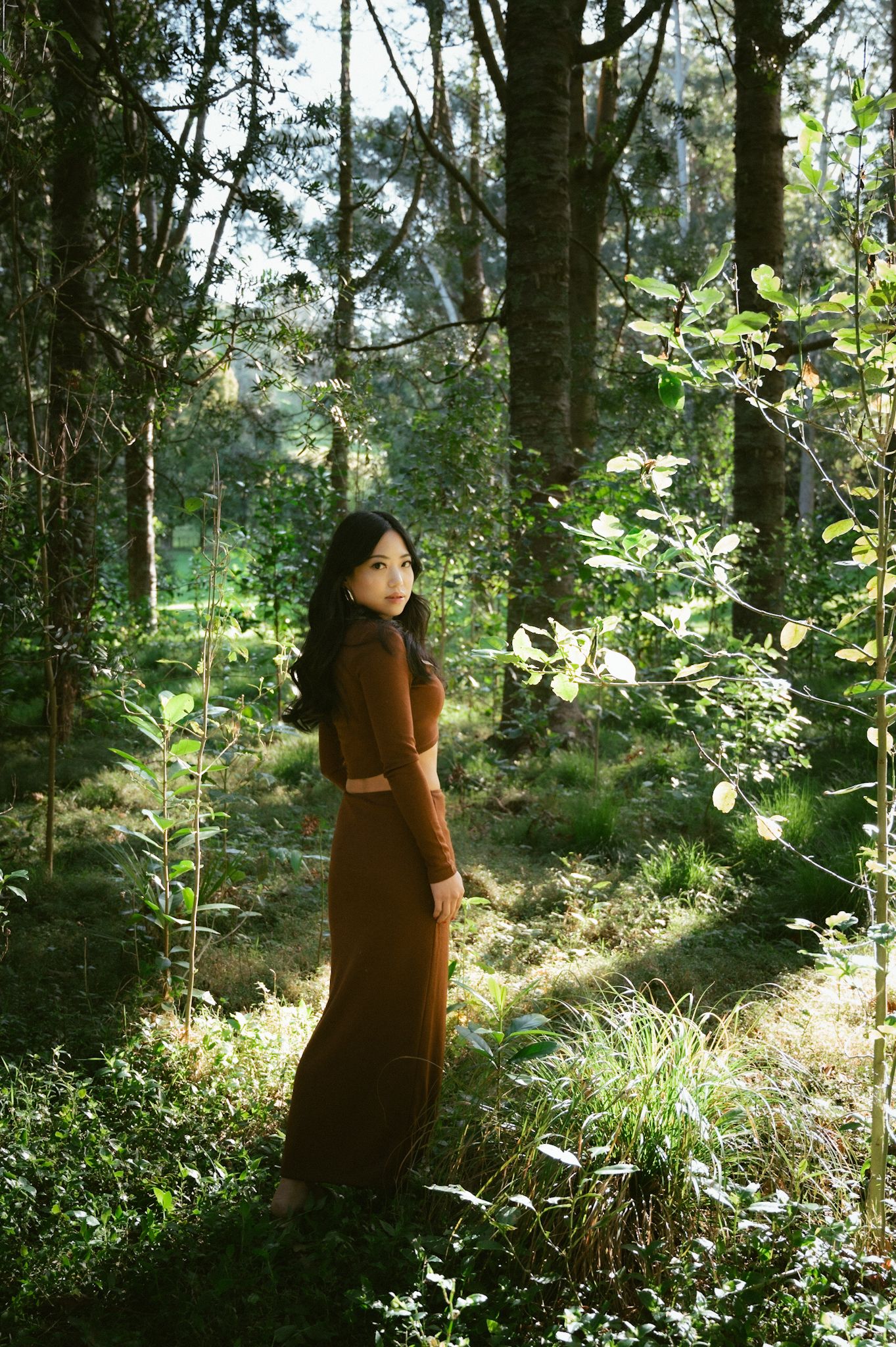 Asian girl in brown dress walking through an ethereal forest with light beaming through.
