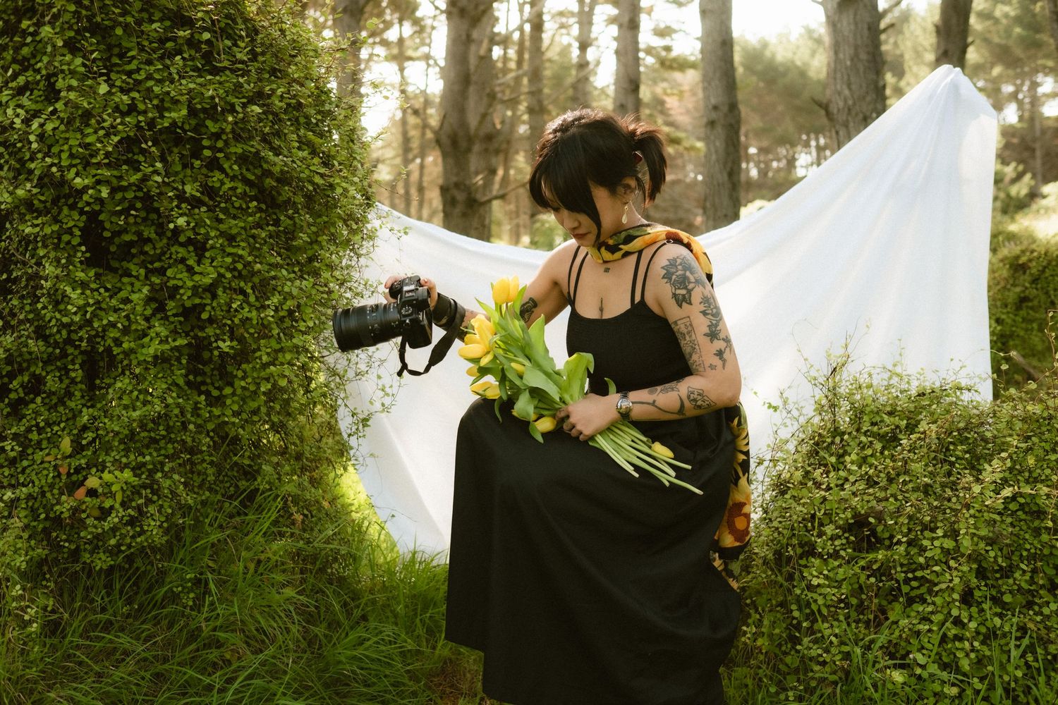Tattooed woman in black dress holding yellow sunflowers and camera outdoors near white fabric backdrop and green hedges.