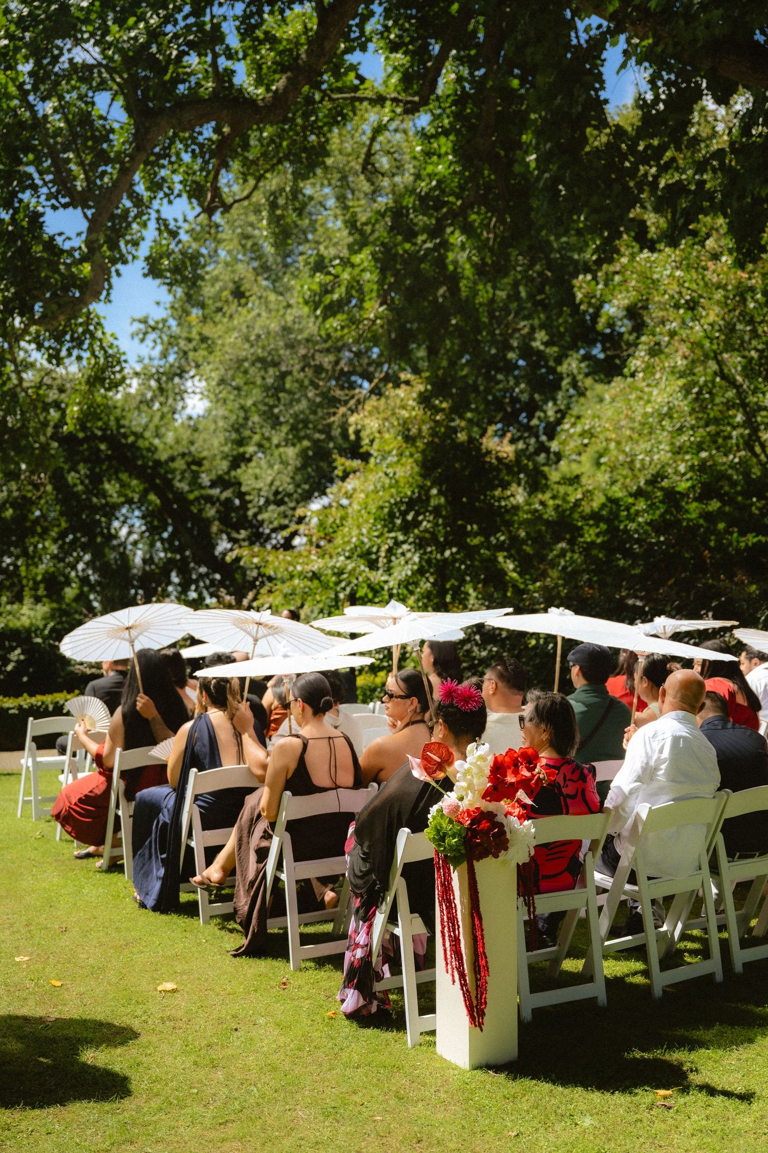 Guests seated in white chairs at an outdoor wedding ceremony under large trees, some holding white parasols on a sunny day.