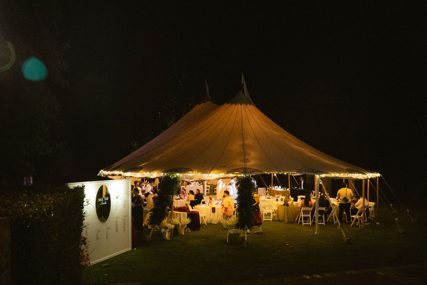 Illuminated wedding tent glowing warmly at night, decorated with string lights and floral arrangements for an outdoor reception.