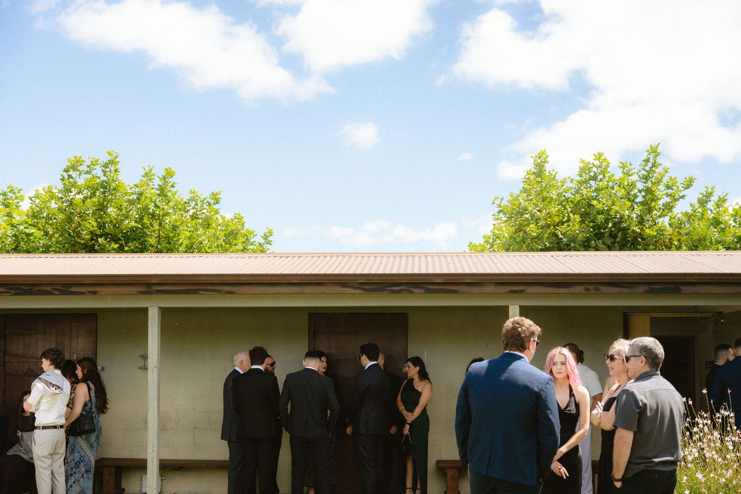 Guests in formal attire mingle outdoors near a low building with green trees under a partly cloudy sky.