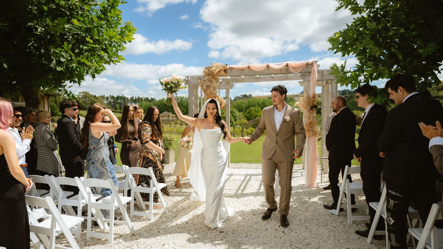 Joyful bride and groom walk down outdoor aisle after wedding ceremony, surrounded by guests under sunny skies.