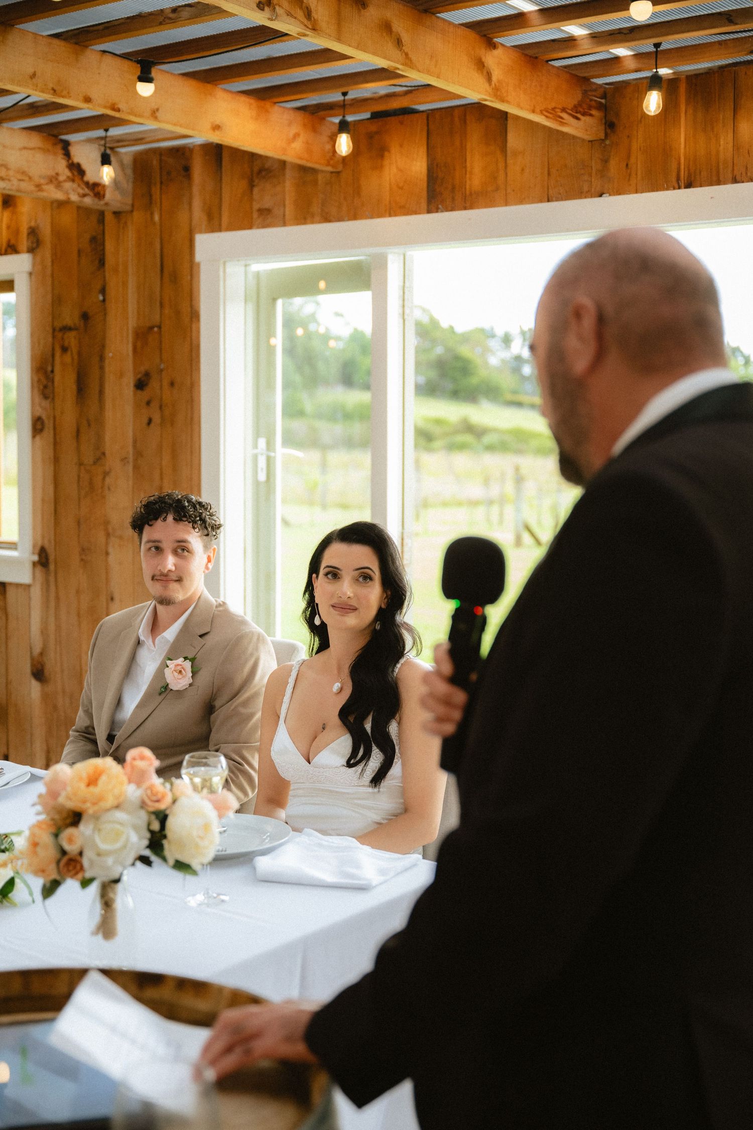 A bald man gives a wedding toast with a microphone as the bride and groom listen attentively at their reception table.