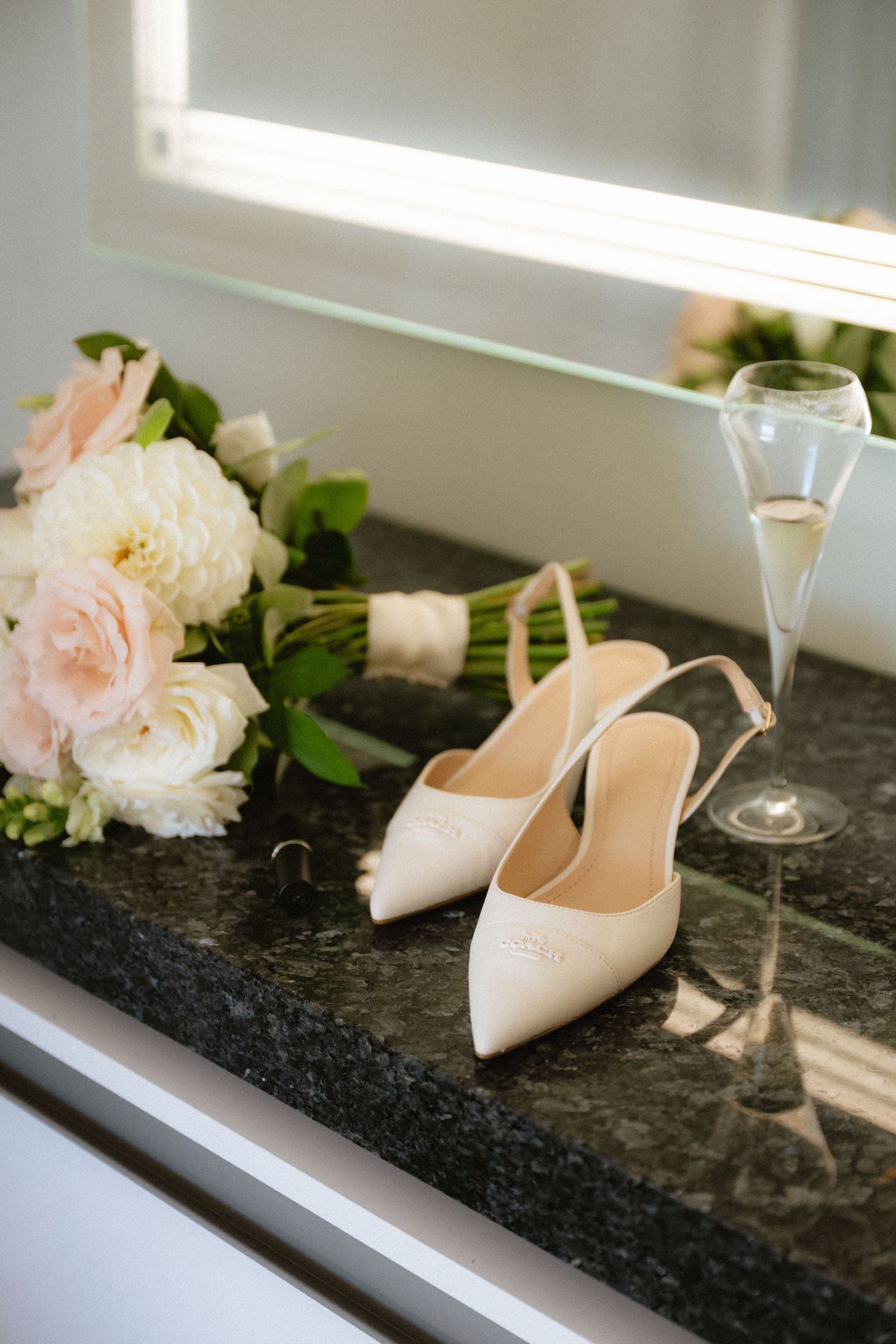 Elegant bridal heels beside a champagne flute and pink and white flower bouquet on a dark marble surface.
