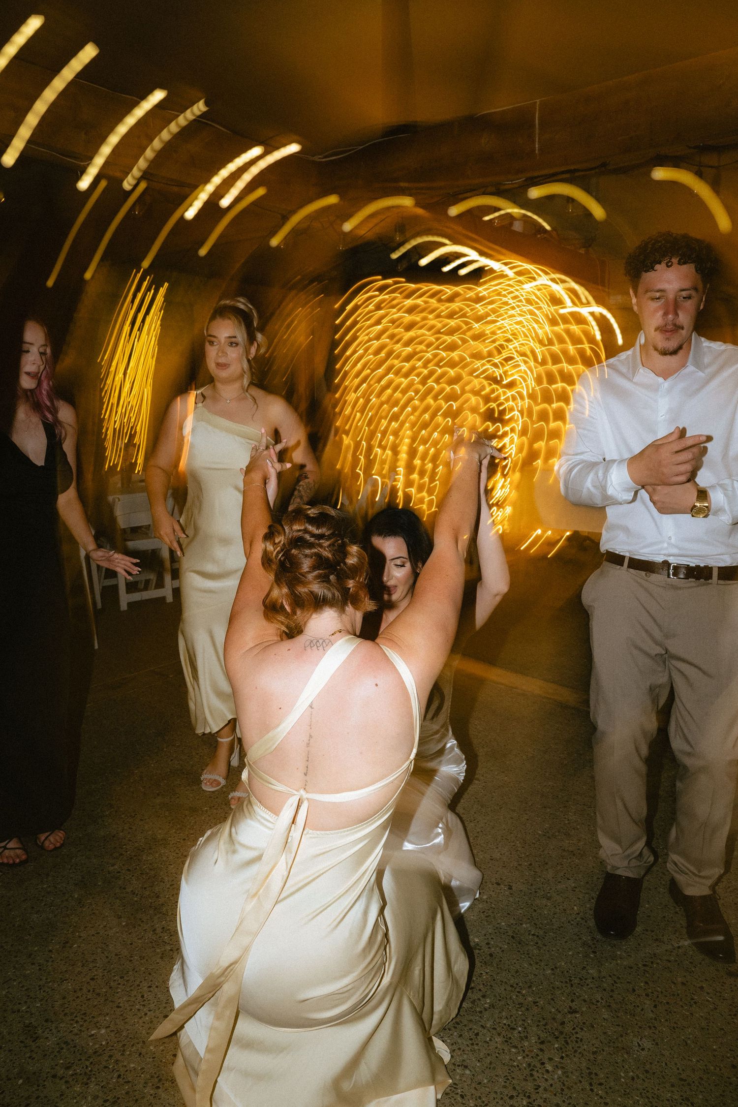 Bridesmaids in gold satin dresses dance joyfully in a warmly lit tunnel venue during a wedding reception.