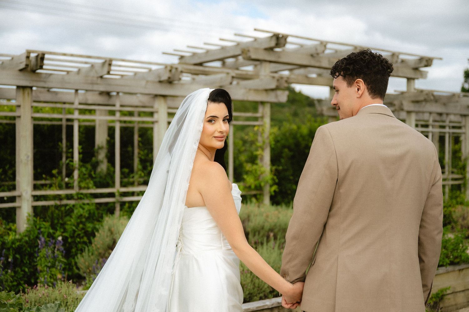 A bride and groom hold hands outdoors at a garden wedding venue, the bride wearing a white gown and veil.