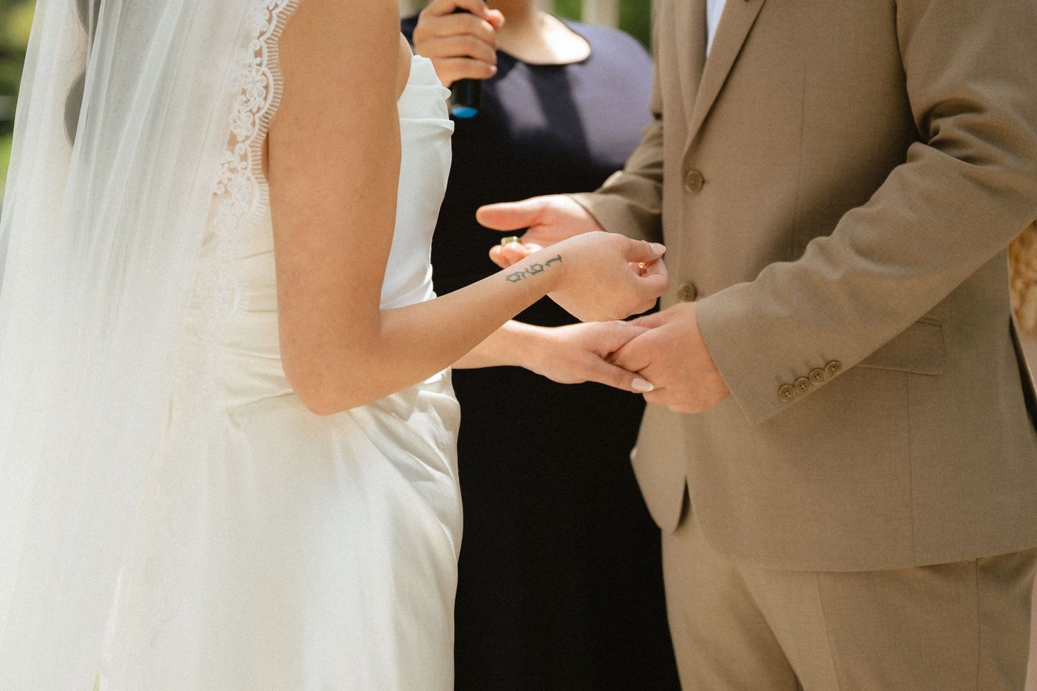 Bride and groom exchanging rings during outdoor wedding ceremony, officiant in background.