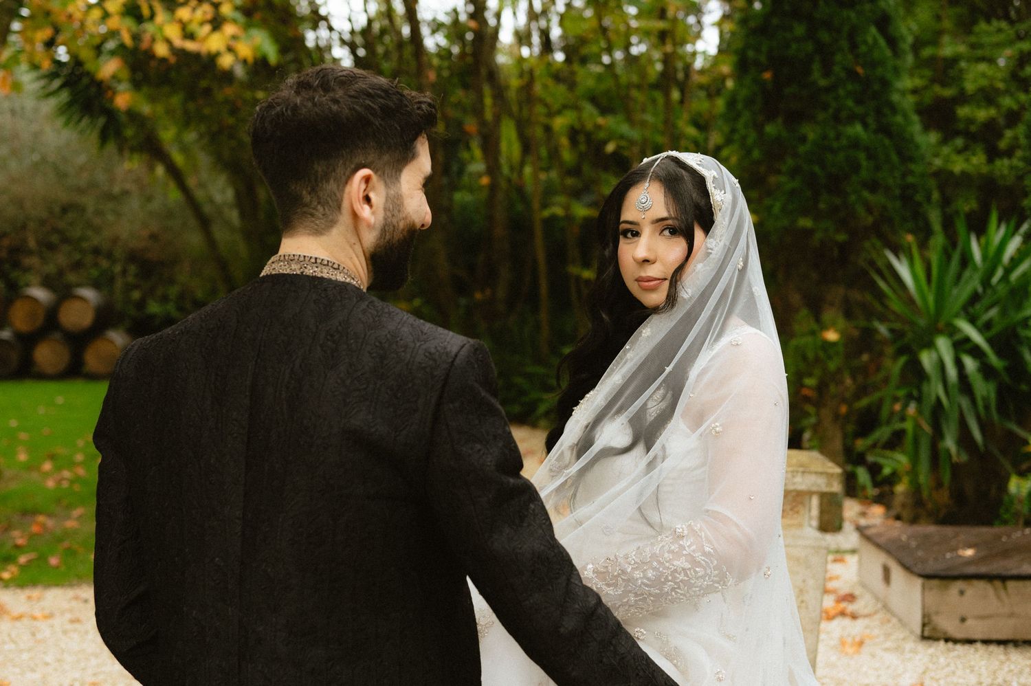 Bride in white veil and groom in black suit hold hands outdoors at an elegant garden wedding ceremony.