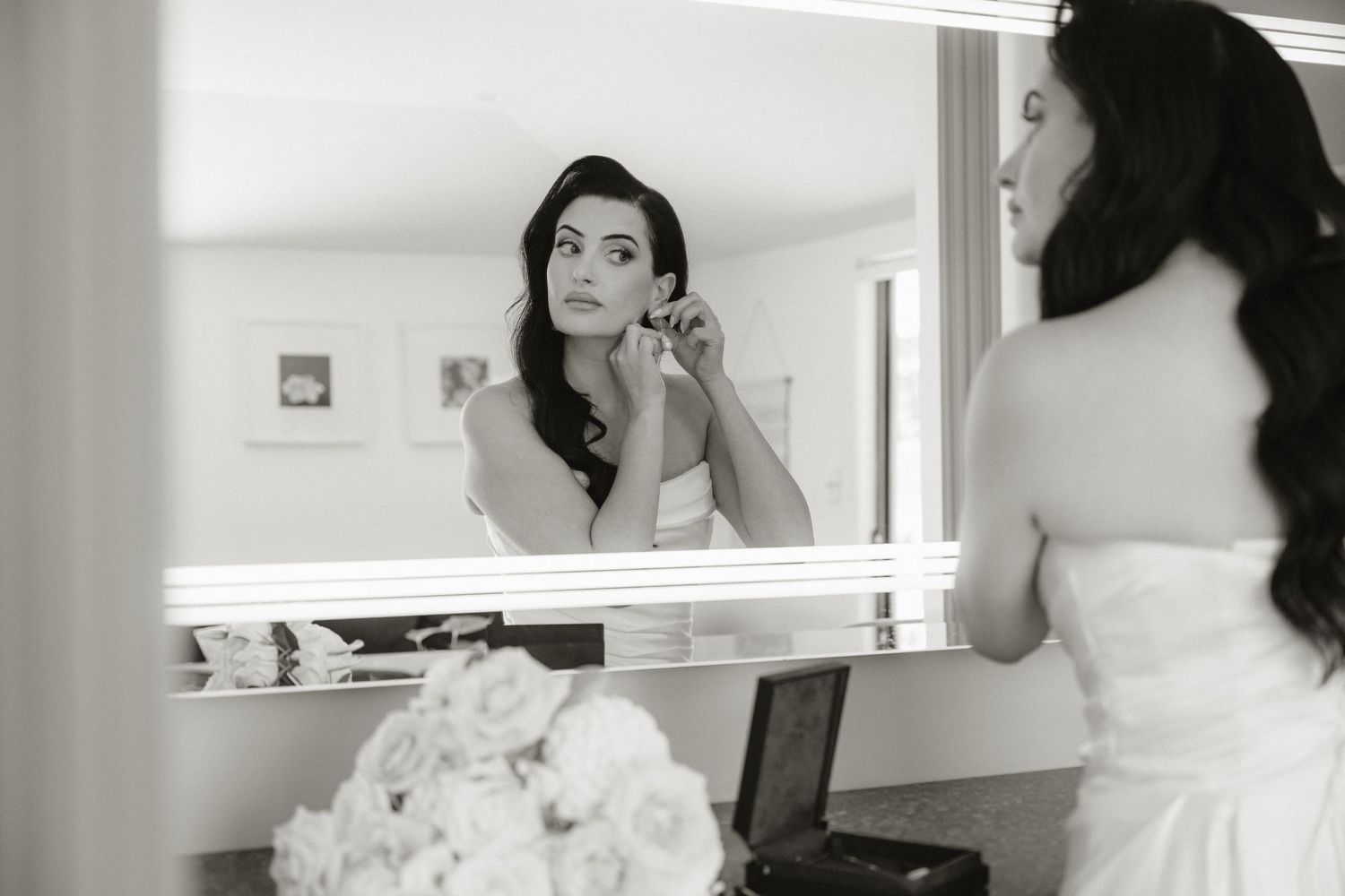 Bride in white gown applying makeup at a lit vanity mirror in a black and white wedding preparation photo.