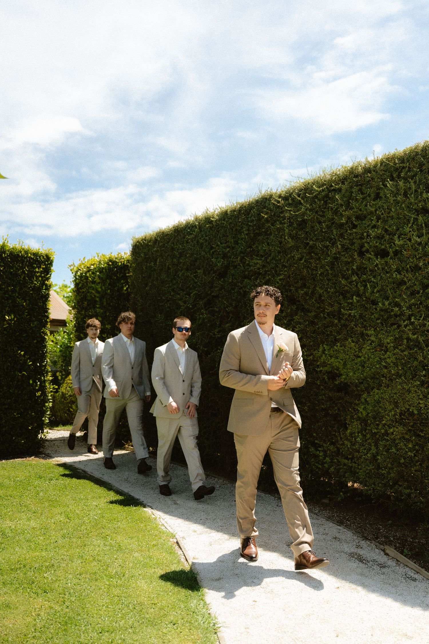 Four groomsmen in tan suits walk along a garden path lined with tall hedges under a bright blue sky.