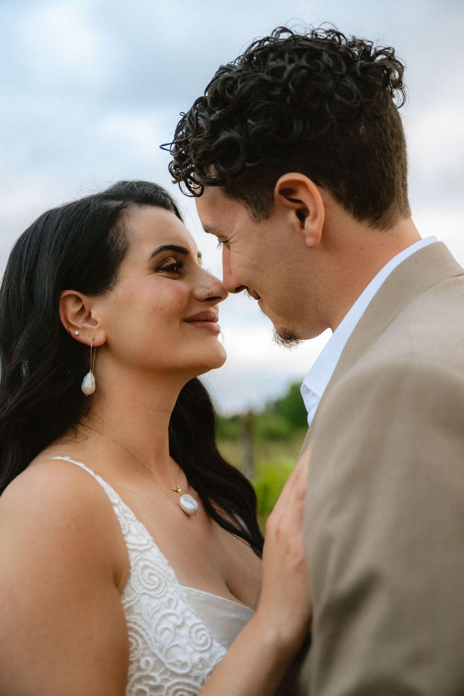 A bride and groom share a romantic close moment, she wearing pearl jewelry and white lace dress, he in a tan suit.