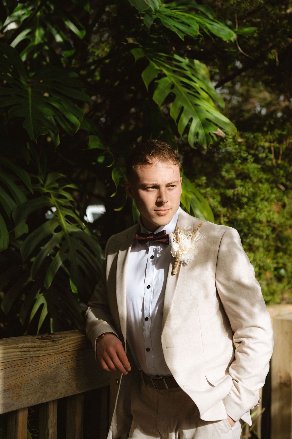 Elegant groom in white tuxedo jacket poses against lush tropical foliage.