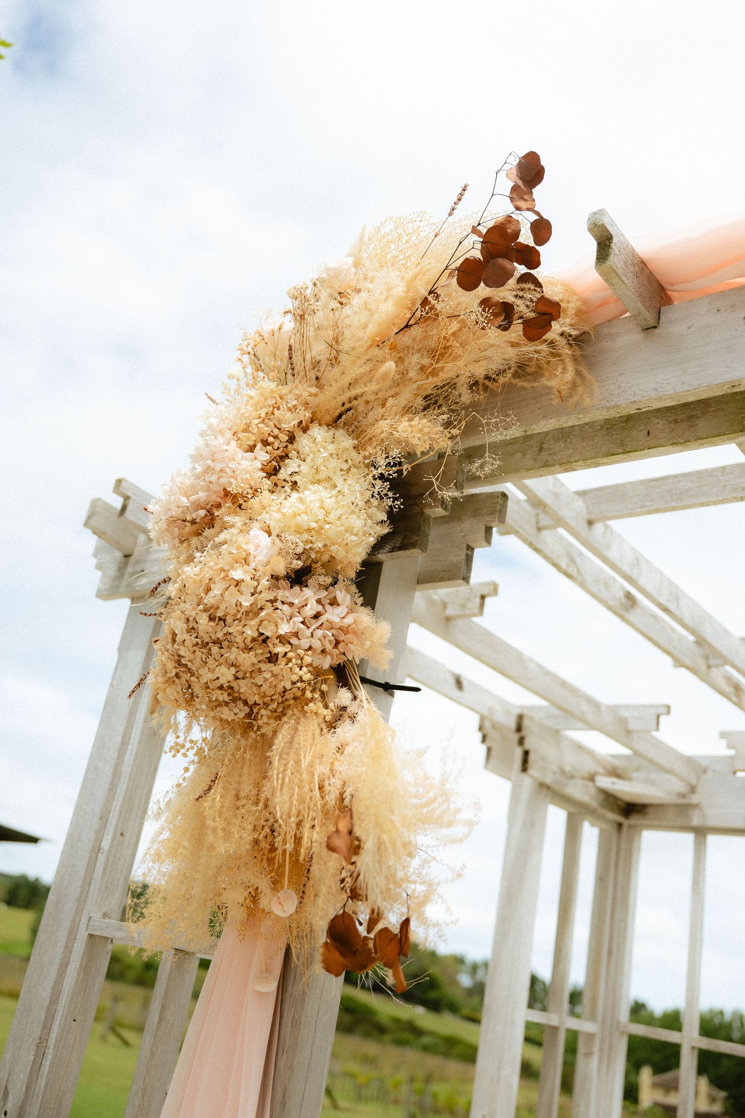 Dried floral wreath with pampas grass and eucalyptus on a white wooden pergola arch outdoors.