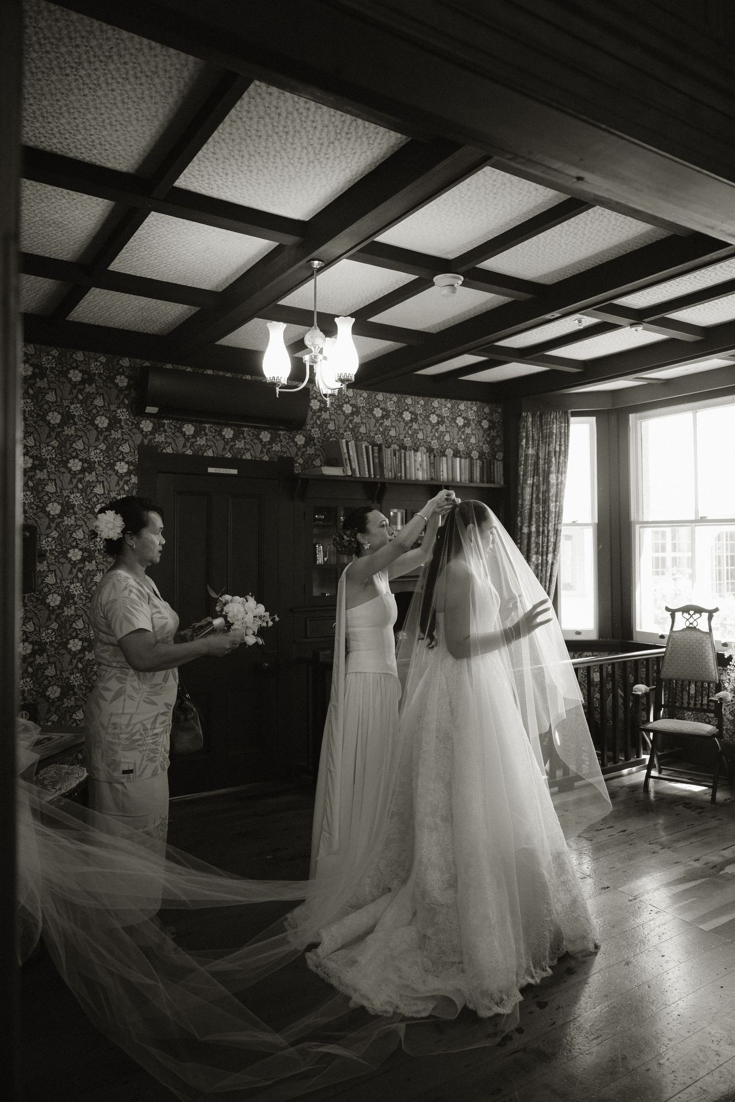 Black and white photo of bride adjusting veil with bridesmaid in elegant wood-paneled room before wedding ceremony.