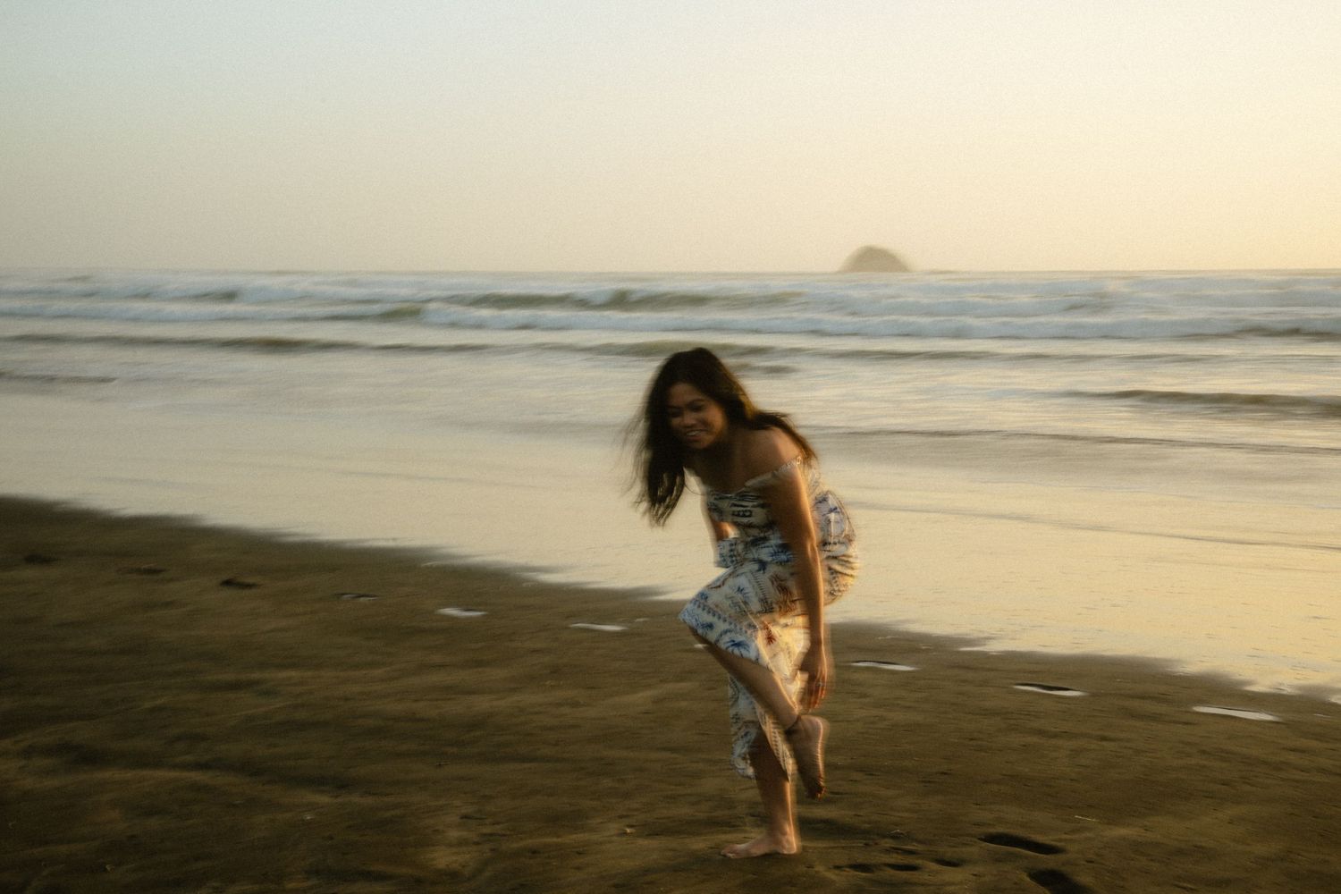 A woman in a floral dress laughs and plays on a sandy beach at sunset with ocean waves behind her.