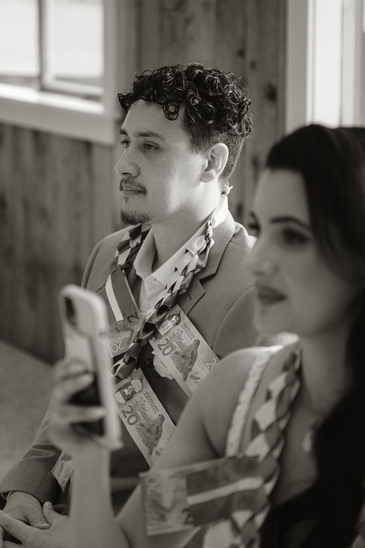 Black and white photo of a man with curly hair holding a phone, with currency pinned to his shirt at a celebration.