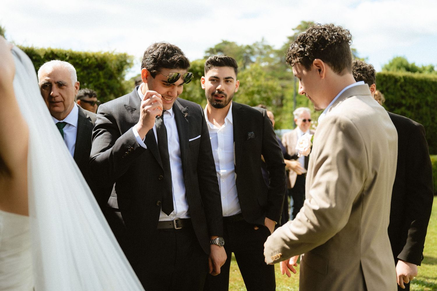 Men in suits conversing outdoors at a wedding reception on a sunny day in a garden setting.
