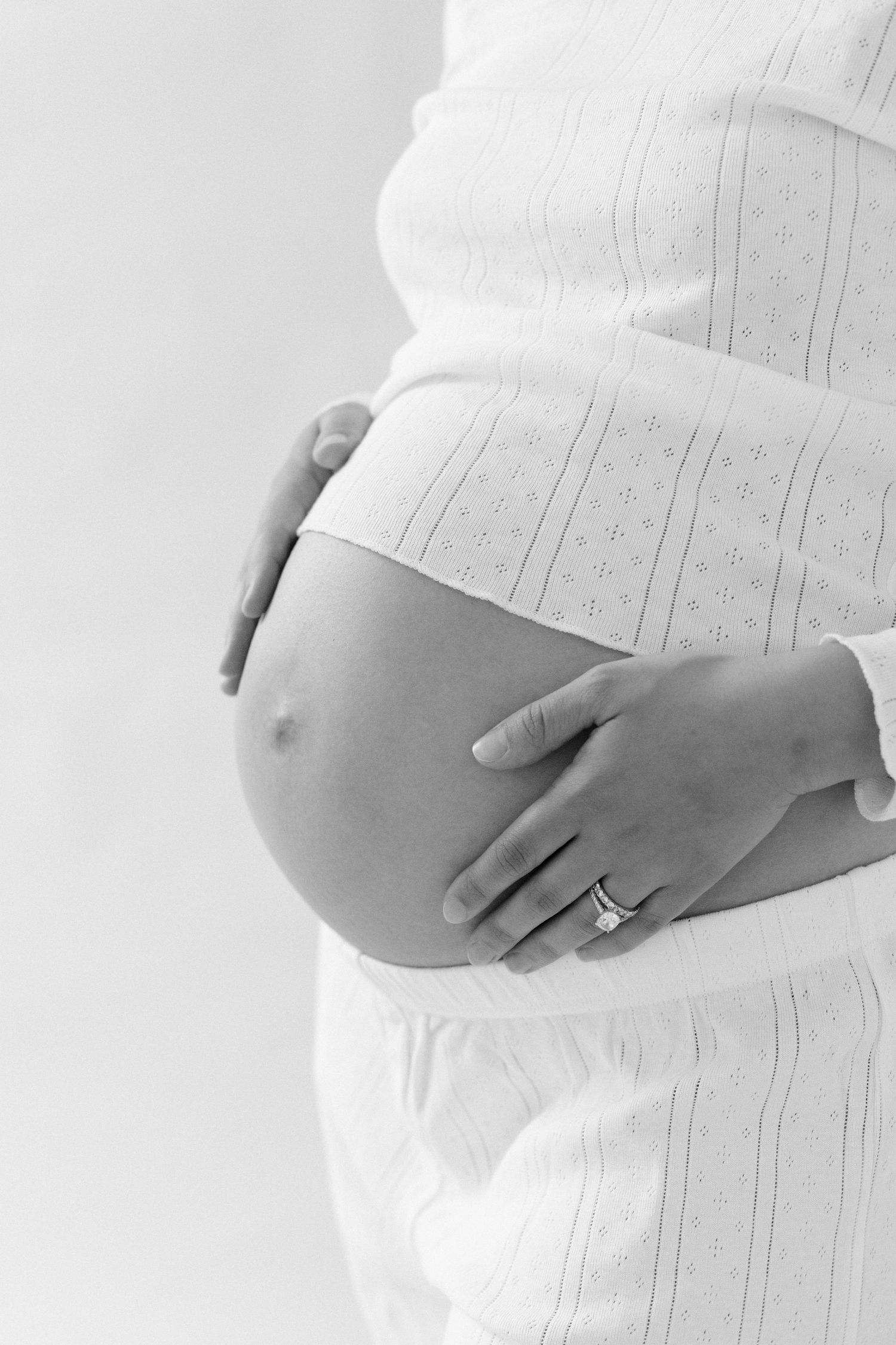 Black and white maternity photo showing a pregnant belly with hands gently resting on it in a white lace dress.