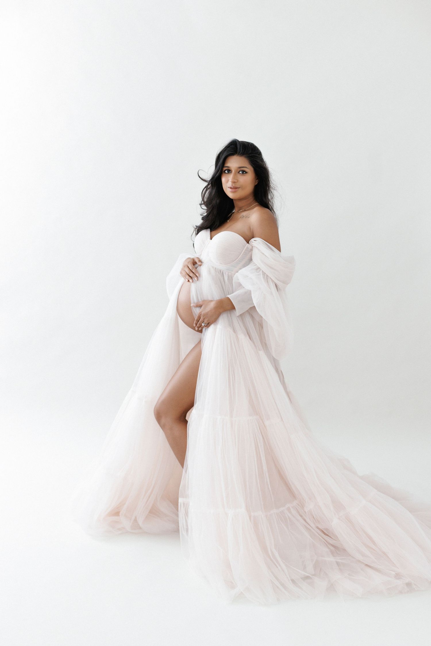 Pregnant woman in elegant white tulle off-shoulder gown cradling baby bump against white background.