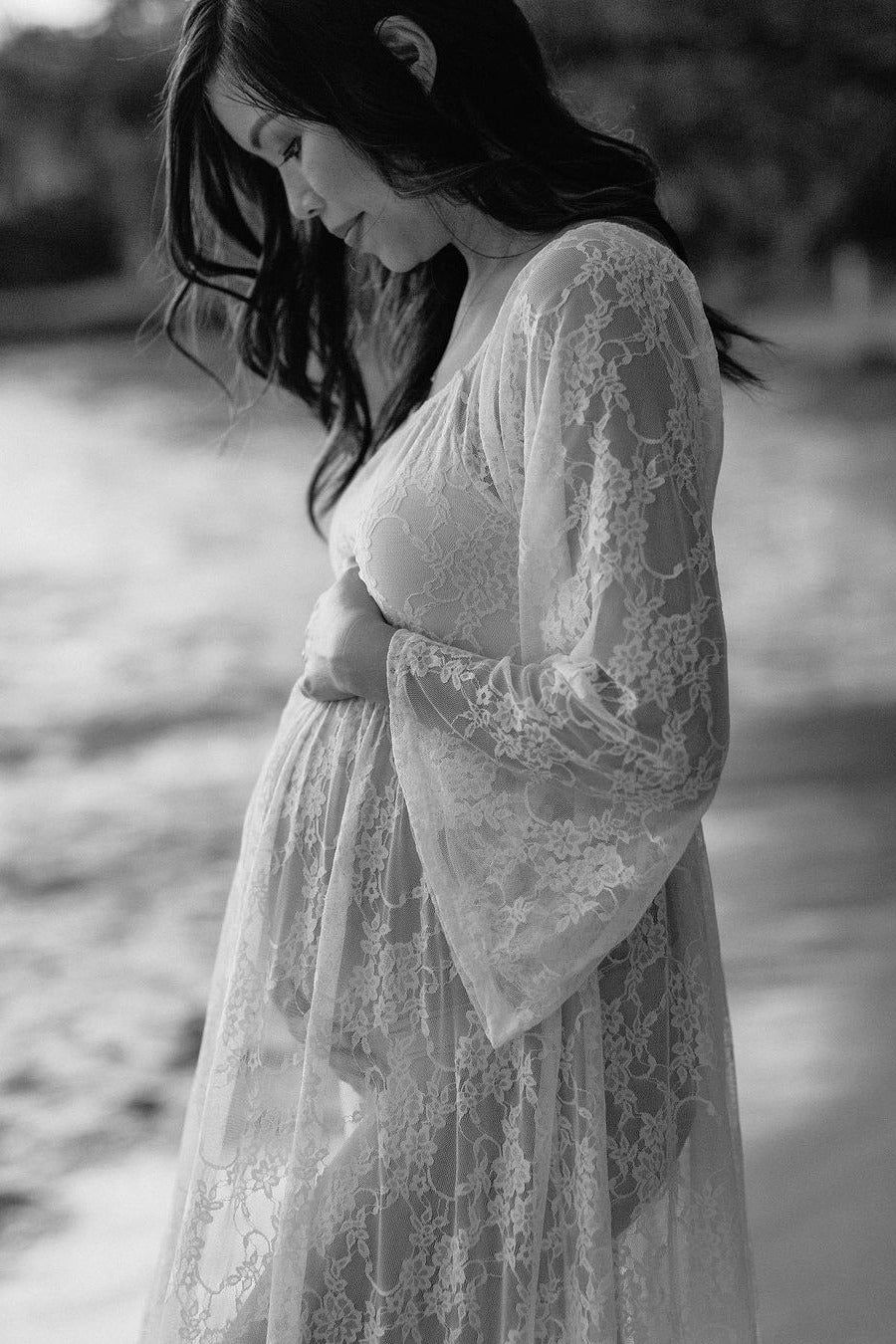 Black and white maternity photo of a woman in a lace dress cradling her baby bump outdoors.