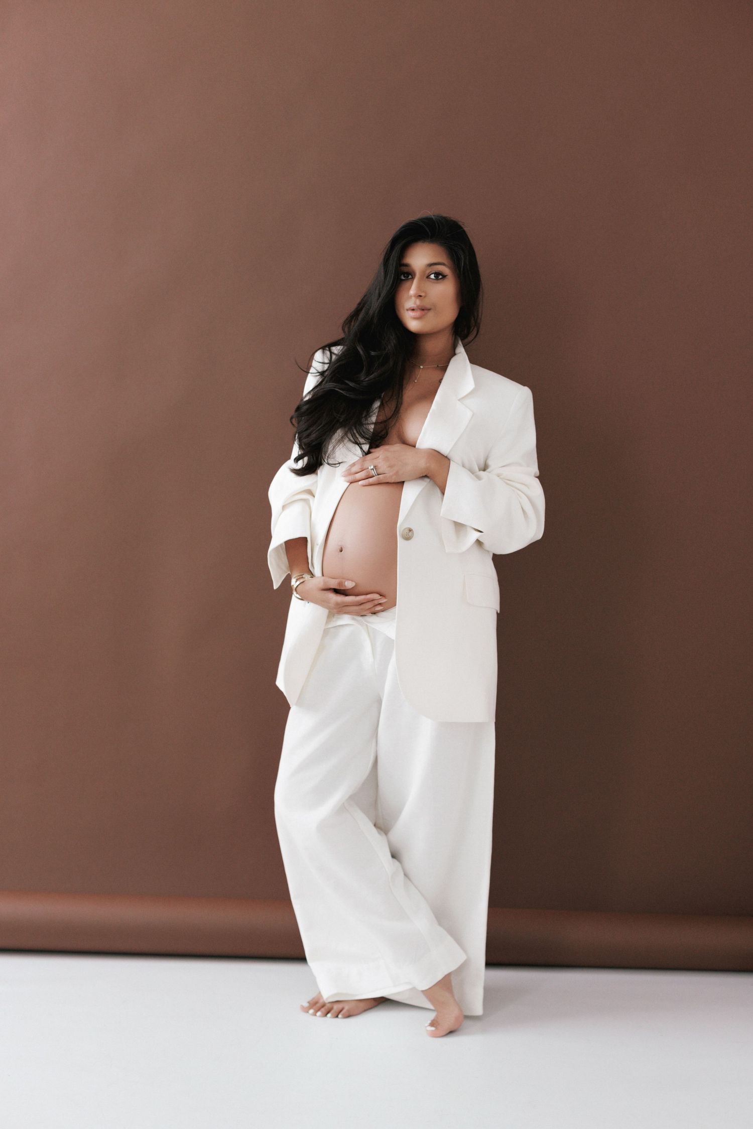 Pregnant woman in white blazer and pants cradling baby bump, posing barefoot against brown studio background.