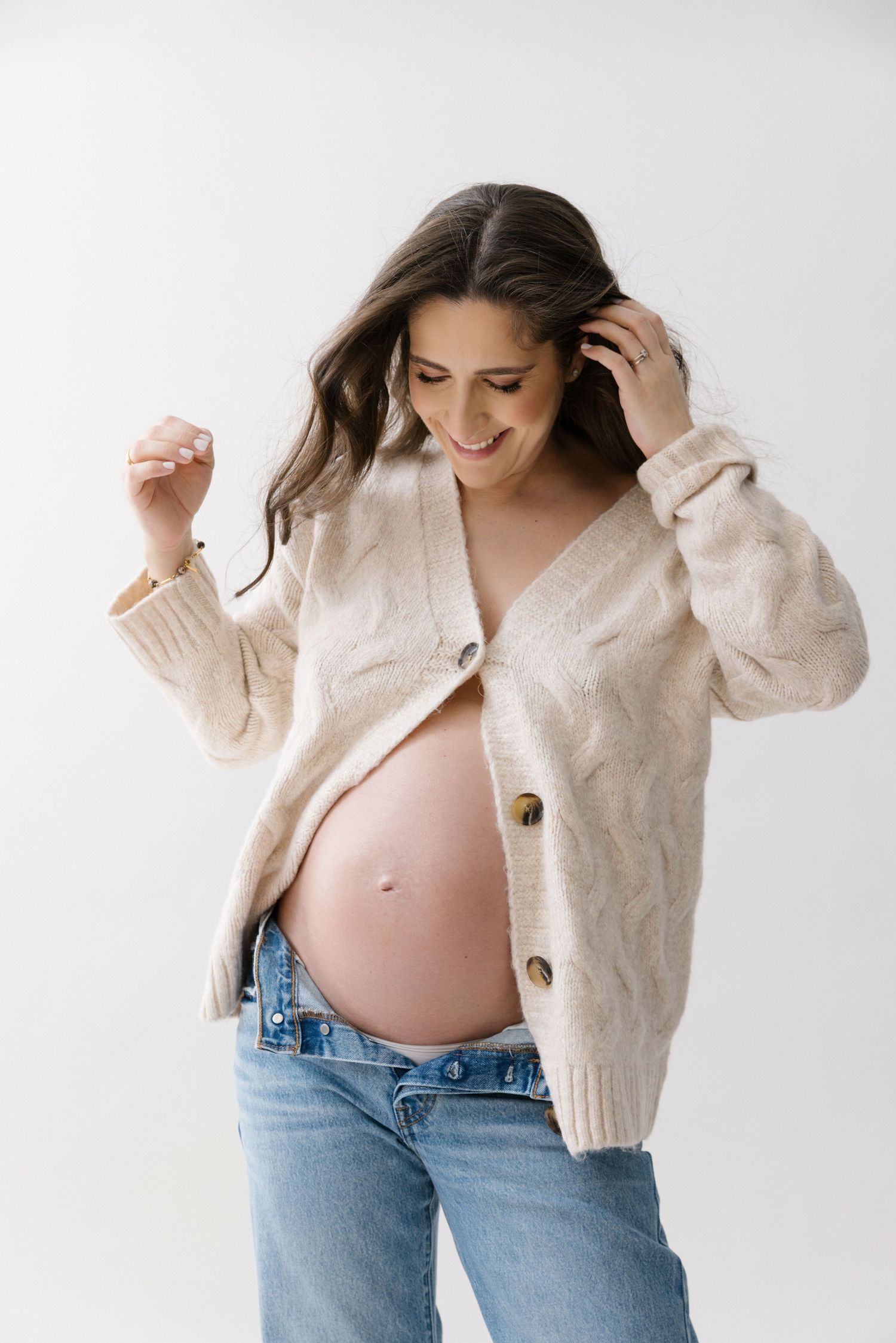 Smiling pregnant woman in beige cardigan and jeans showing baby bump, posing against white background.