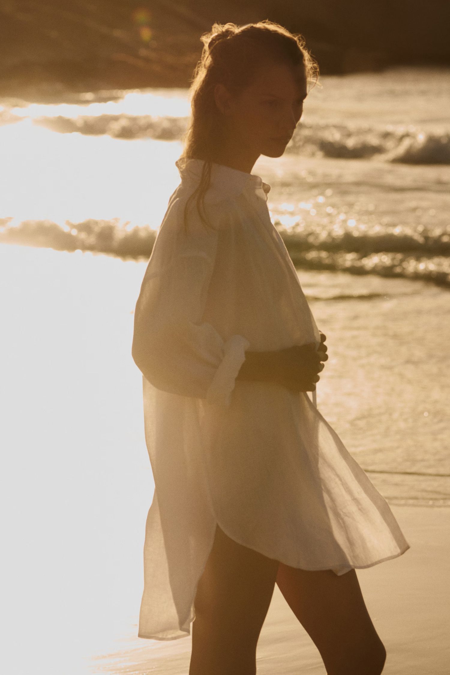 Woman in sheer white shirt walking on beach during golden sunset.